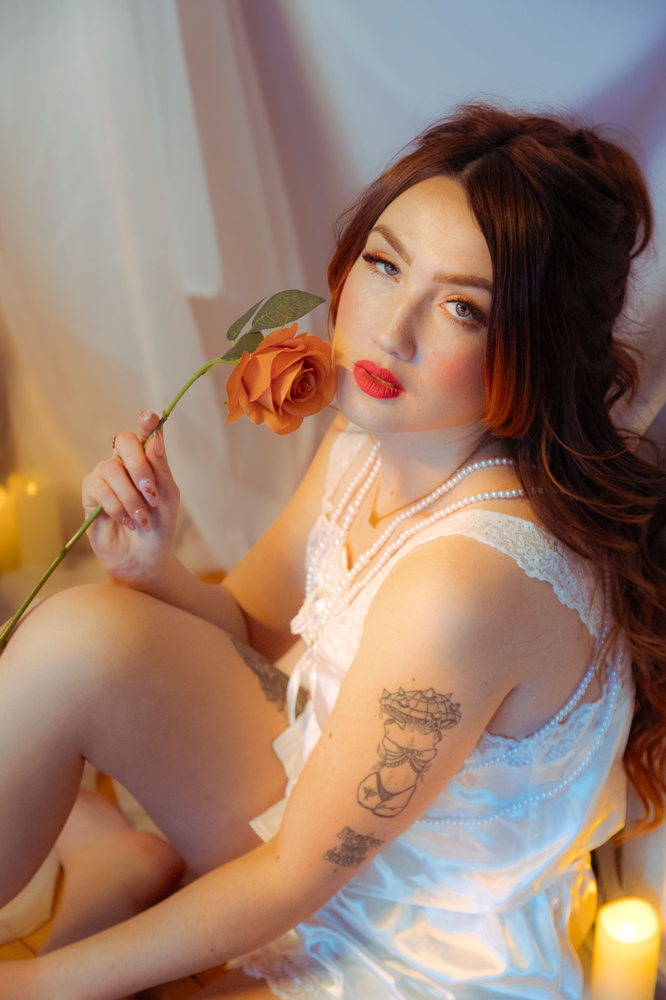 A woman in white lace lingerie holds an orange rose, adorned with pearls, surrounded by candles in a romantic setting.