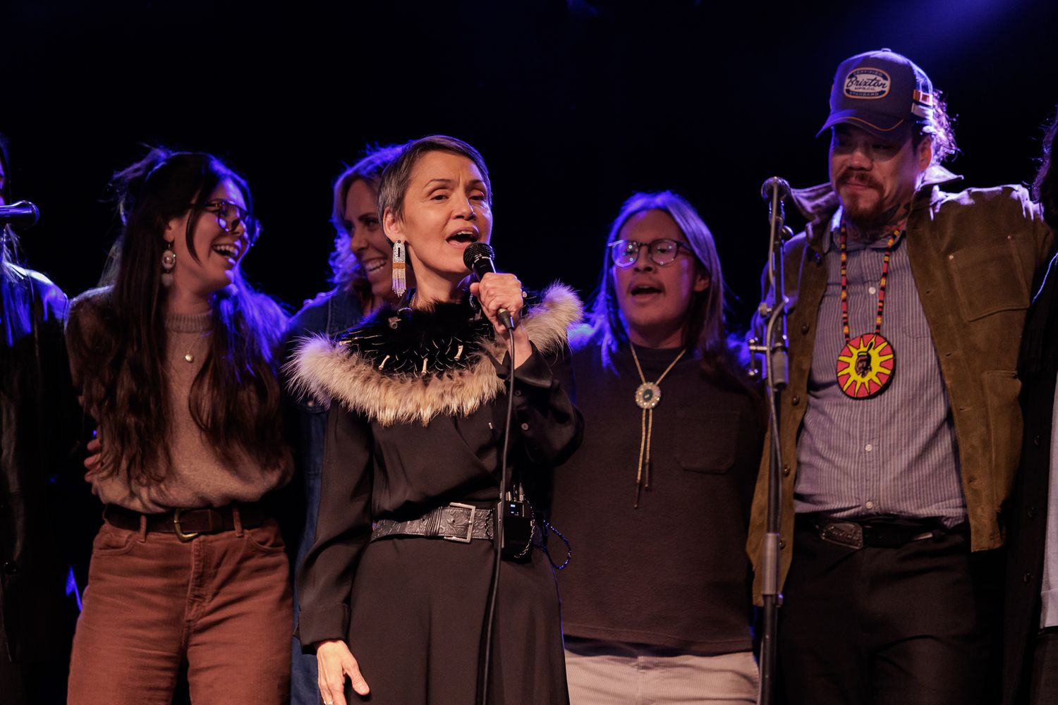 A group of five performers smile and sing together on stage under colorful lights at a live music event.
