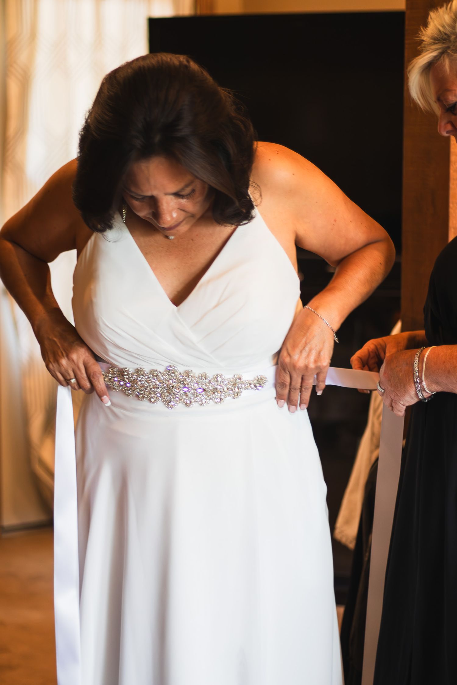 Bride in white V-neck gown with rhinestone belt being helped with dress by assistant before wedding ceremony.