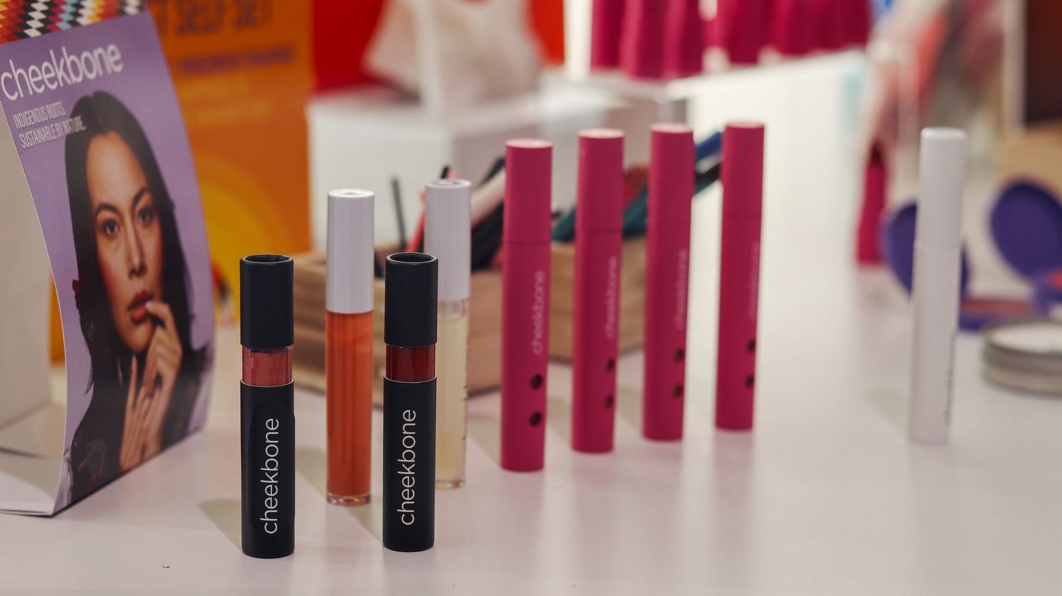 Cheekbone Beauty lip gloss and lipstick products displayed on a white table at a cosmetics showcase event.