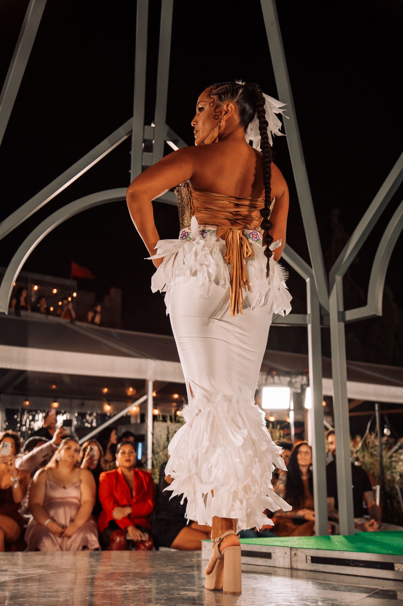 Model in white feathered mermaid gown with brown corset back walks runway at night fashion show.