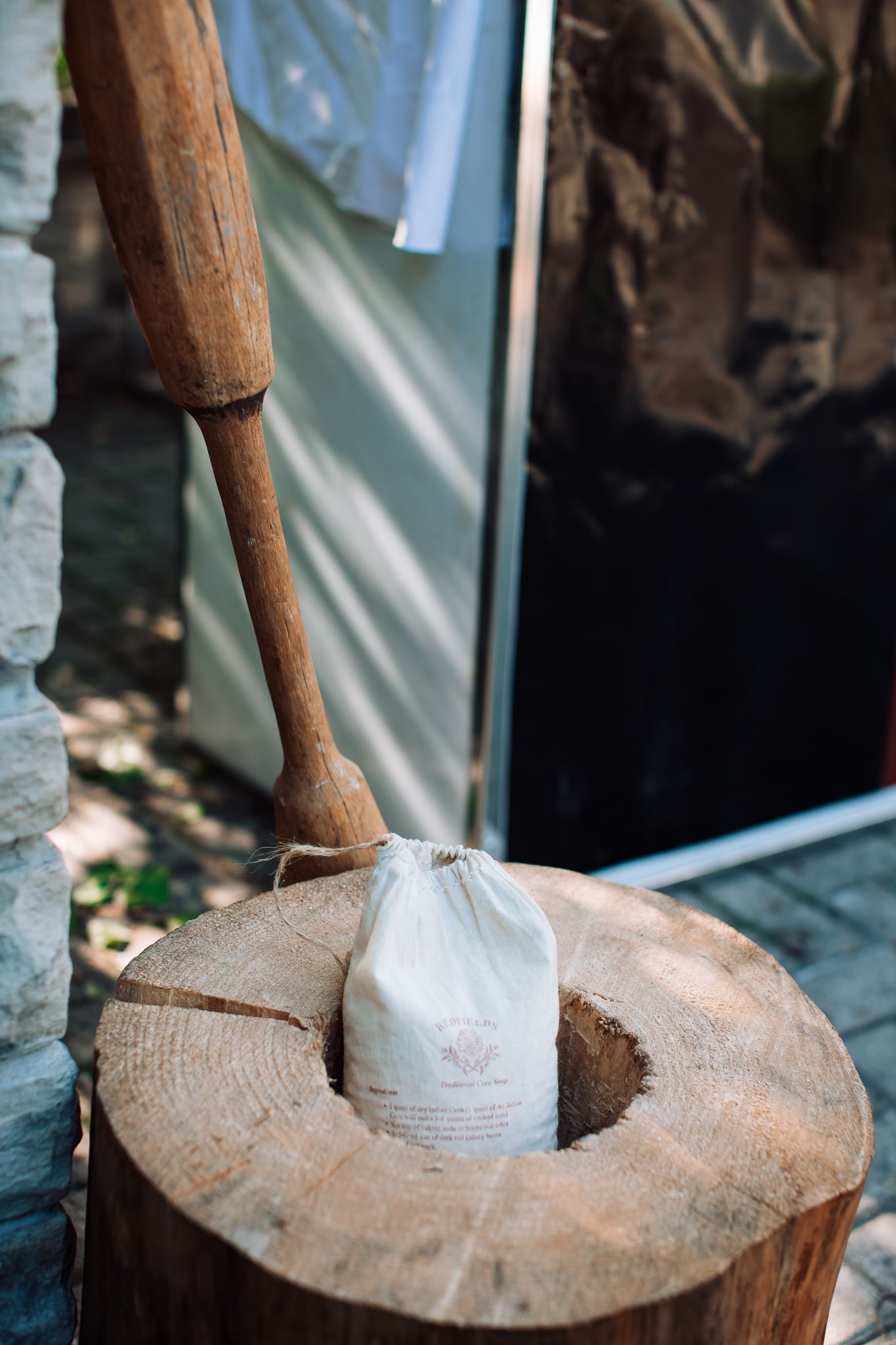 Wooden pestle resting on a carved tree stump with a small white linen drawstring bag in an outdoor setting.