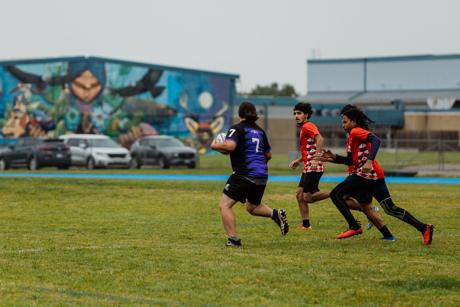 Students play flag football on a grass field with a colorful mural and parked cars in the background.