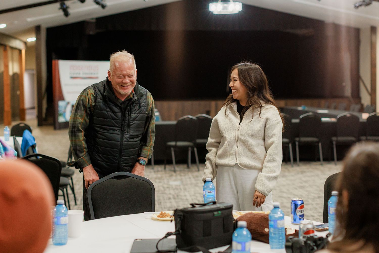 Two people standing and conversing at a conference room event with water bottles and chairs in the background.