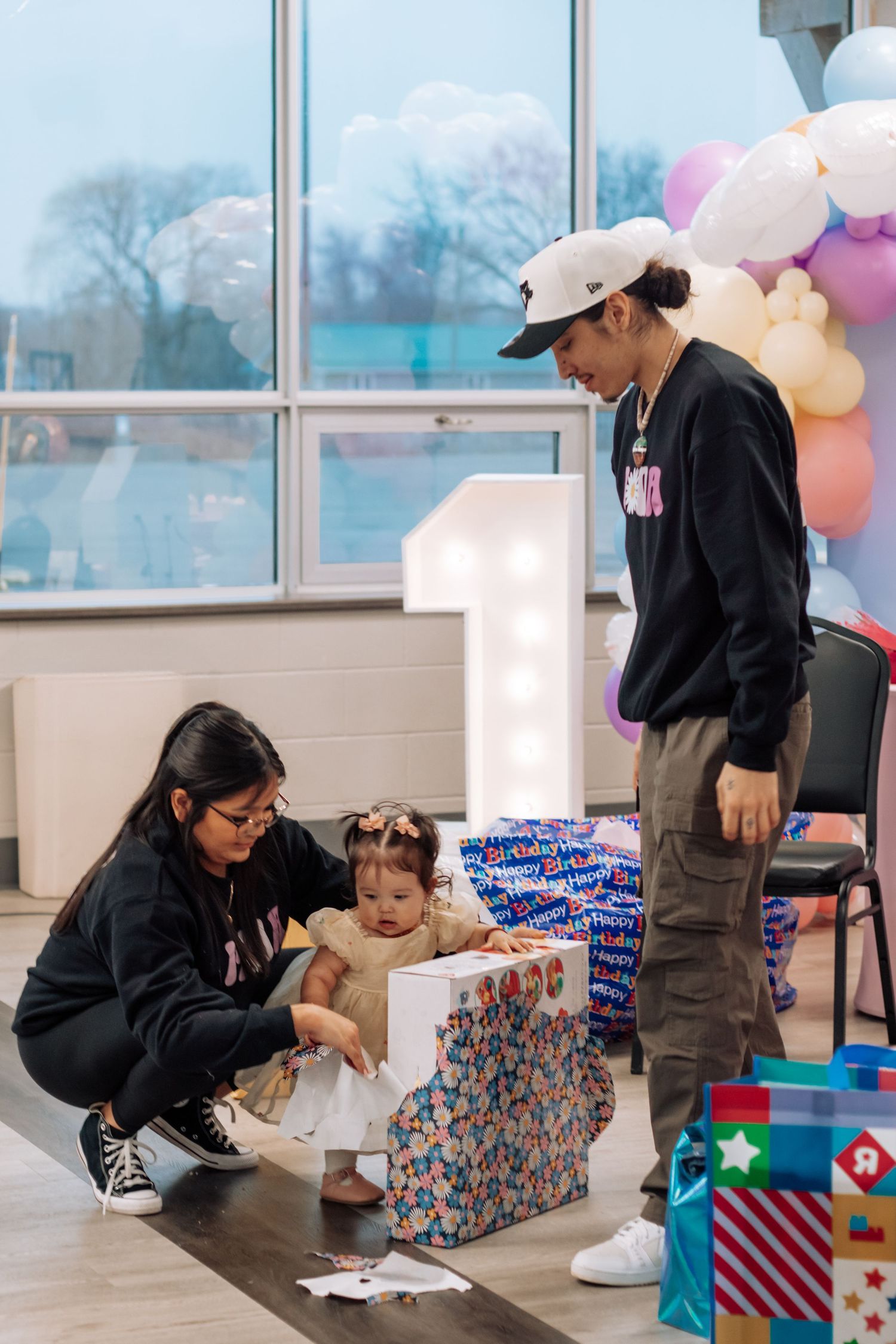 Family celebrating baby's first birthday party with balloons and gifts in a decorated event venue.