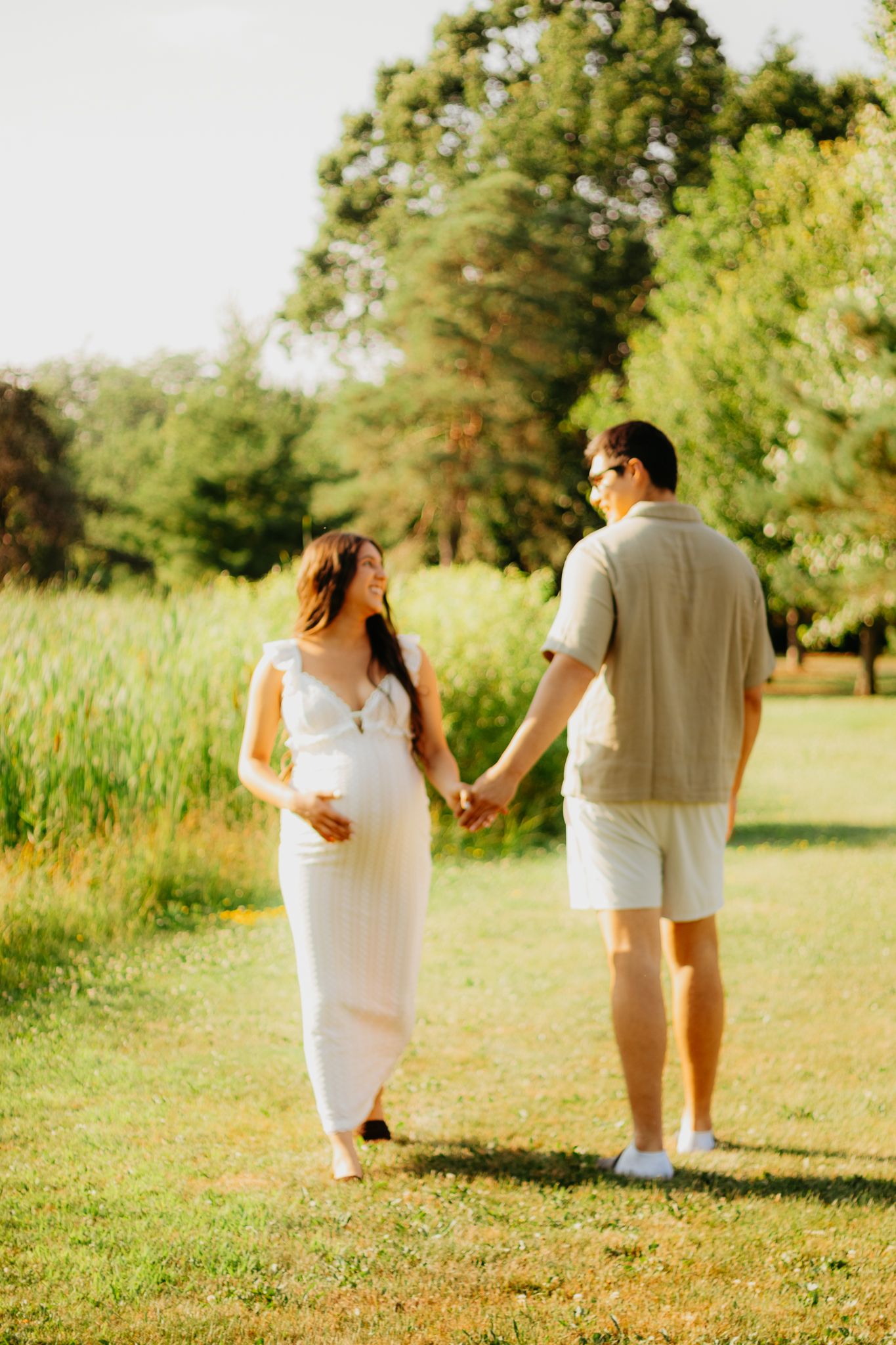 Candid walking shots of an expectant couple holding hands in a sun-filled meadow.