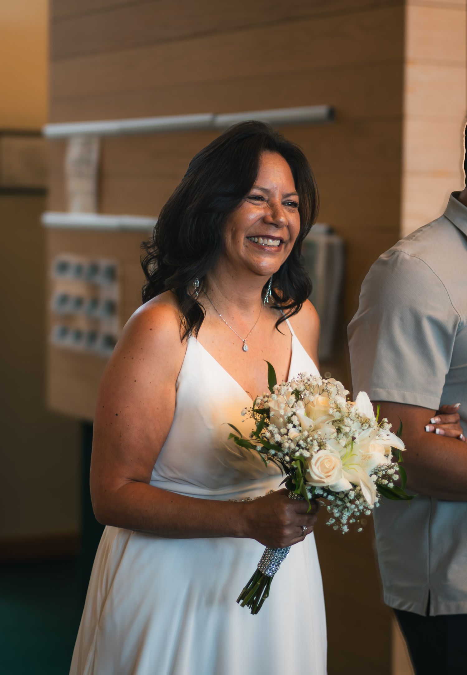 Smiling bride in white spaghetti strap gown holding white rose and baby's breath bouquet at wedding.