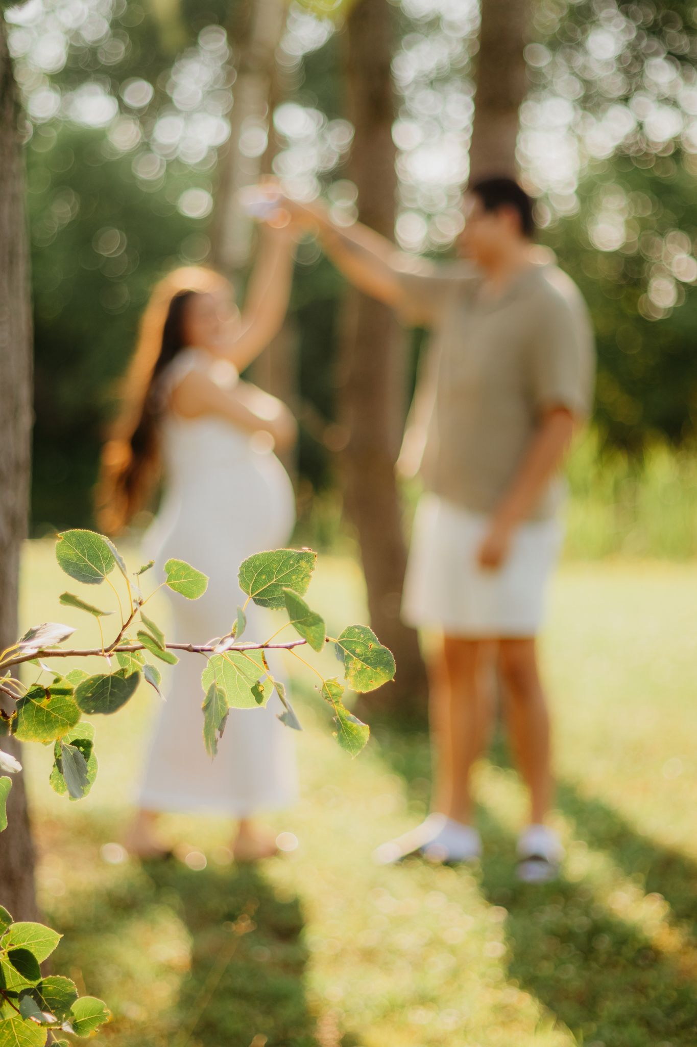 Blurred figures in white clothing dance in a sunlit forest with green foliage in the foreground.
