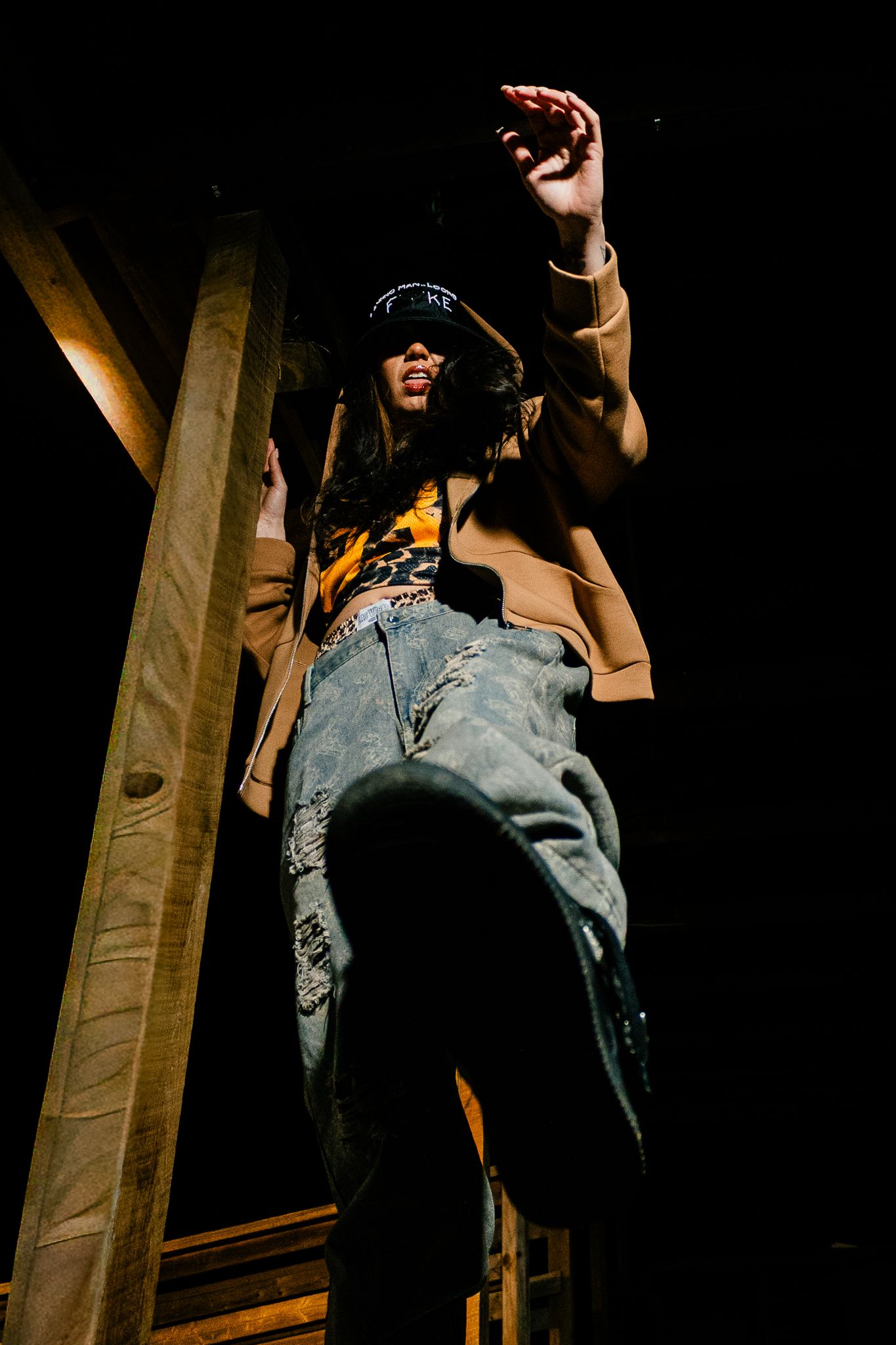 Person in bucket hat, tan jacket, distressed jeans posing dramatically at night near wooden structure.