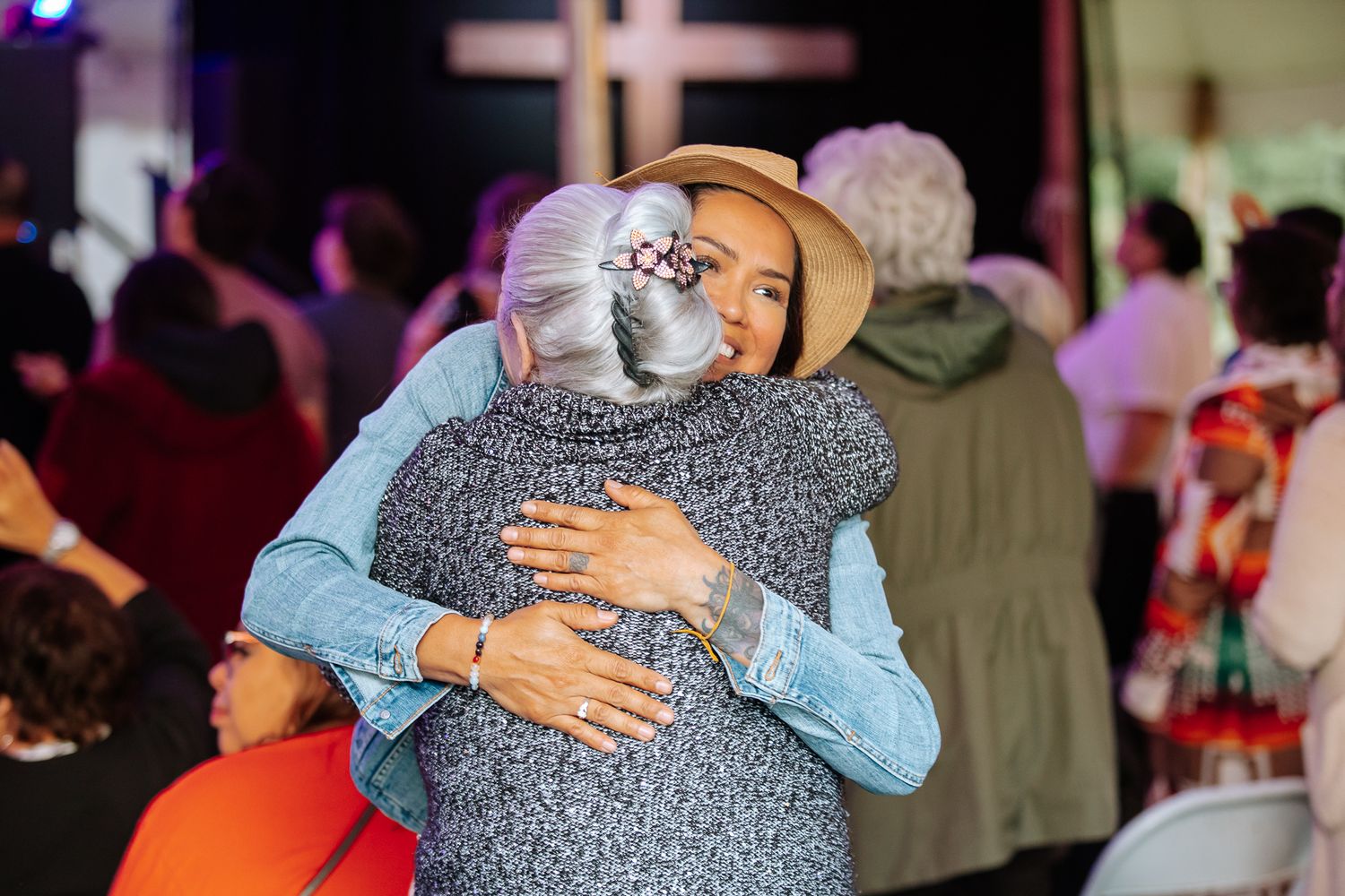 Two women share a warm embrace at a festive indoor gathering with colorful decorations and a cross in the background.