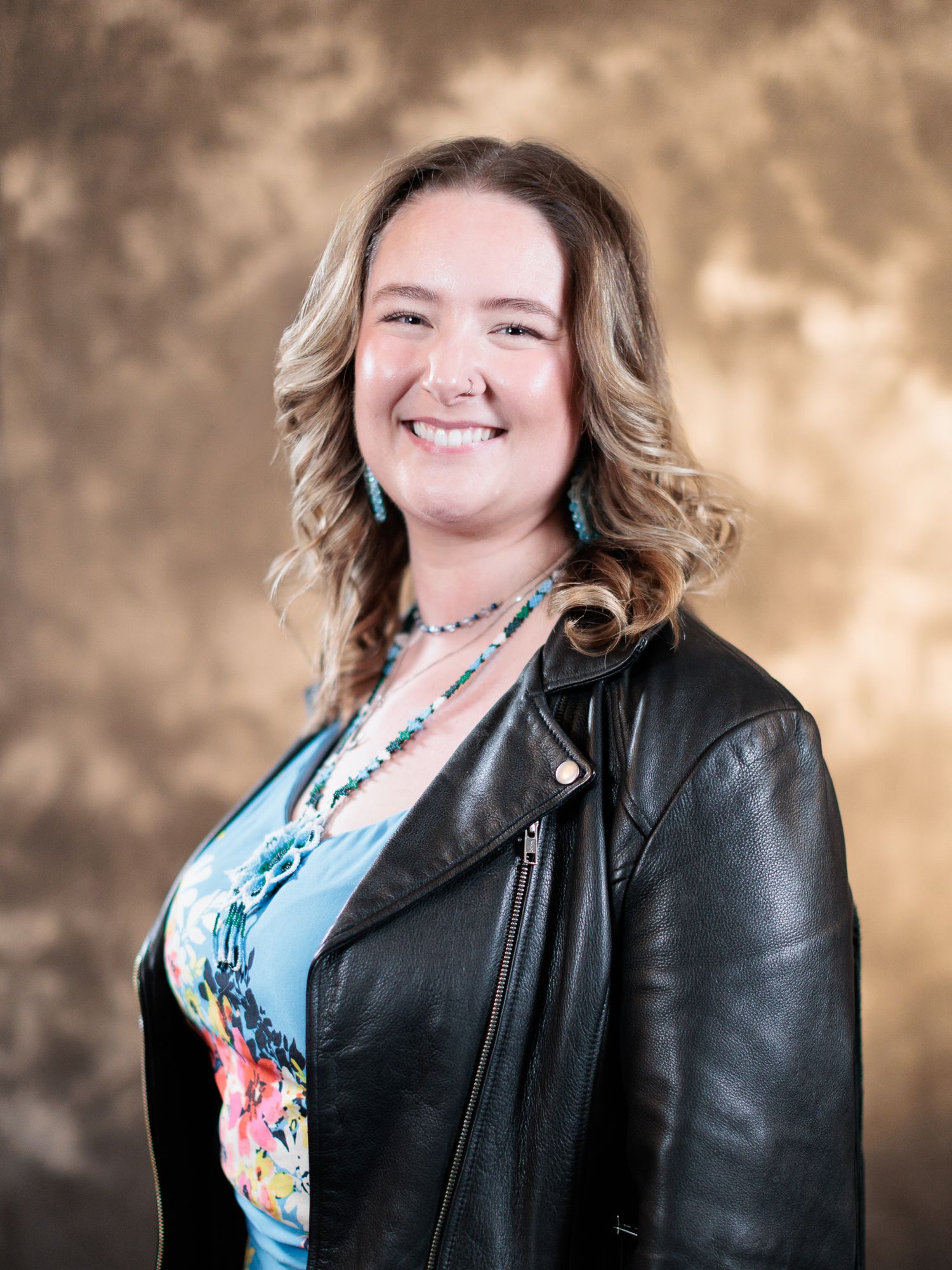 Smiling woman with curly blonde hair wearing a black leather jacket and turquoise jewelry poses for a professional portrait.