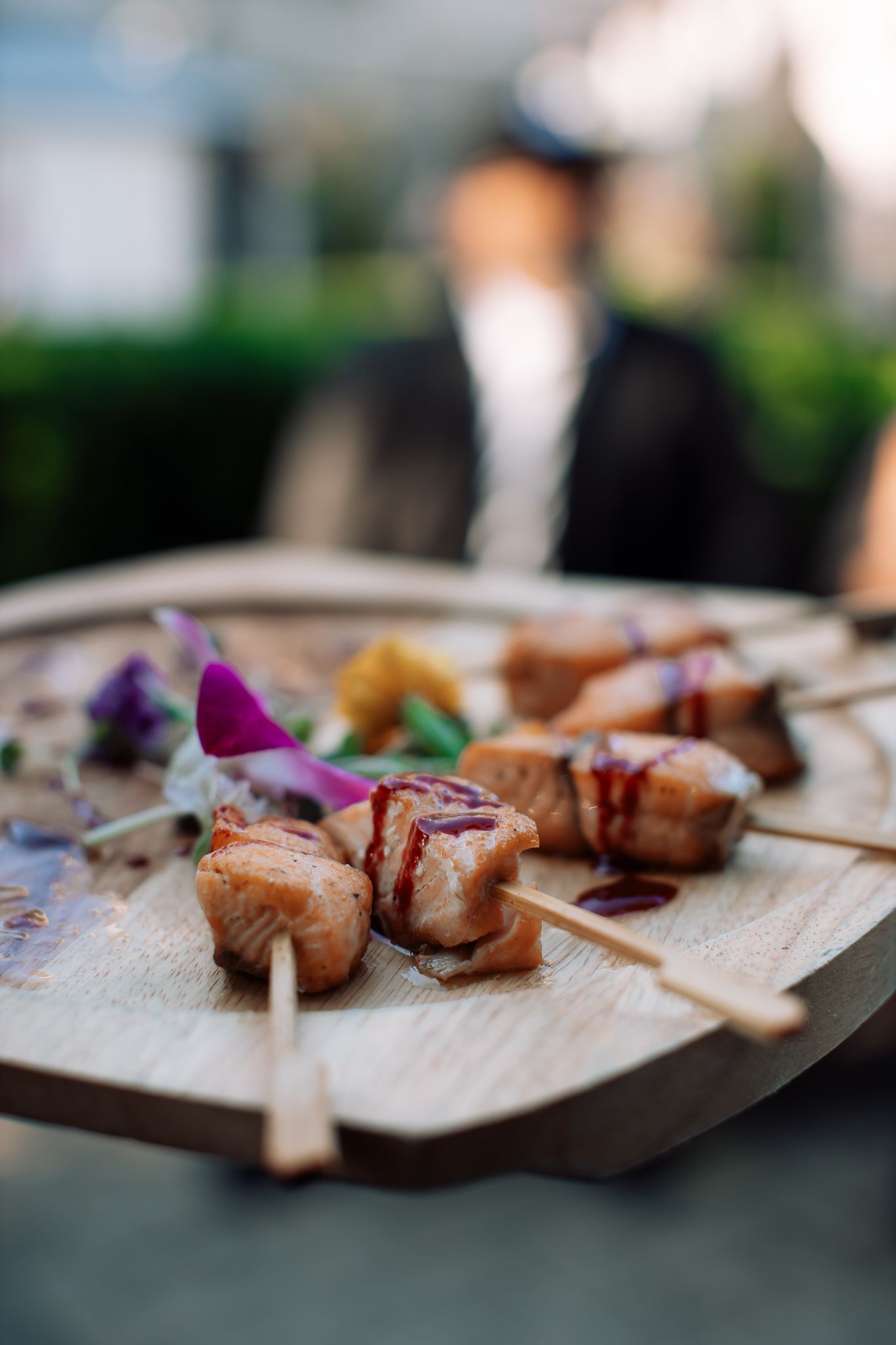 Grilled salmon skewers garnished with edible flowers and sauce served on a rustic wooden board at an outdoor event.