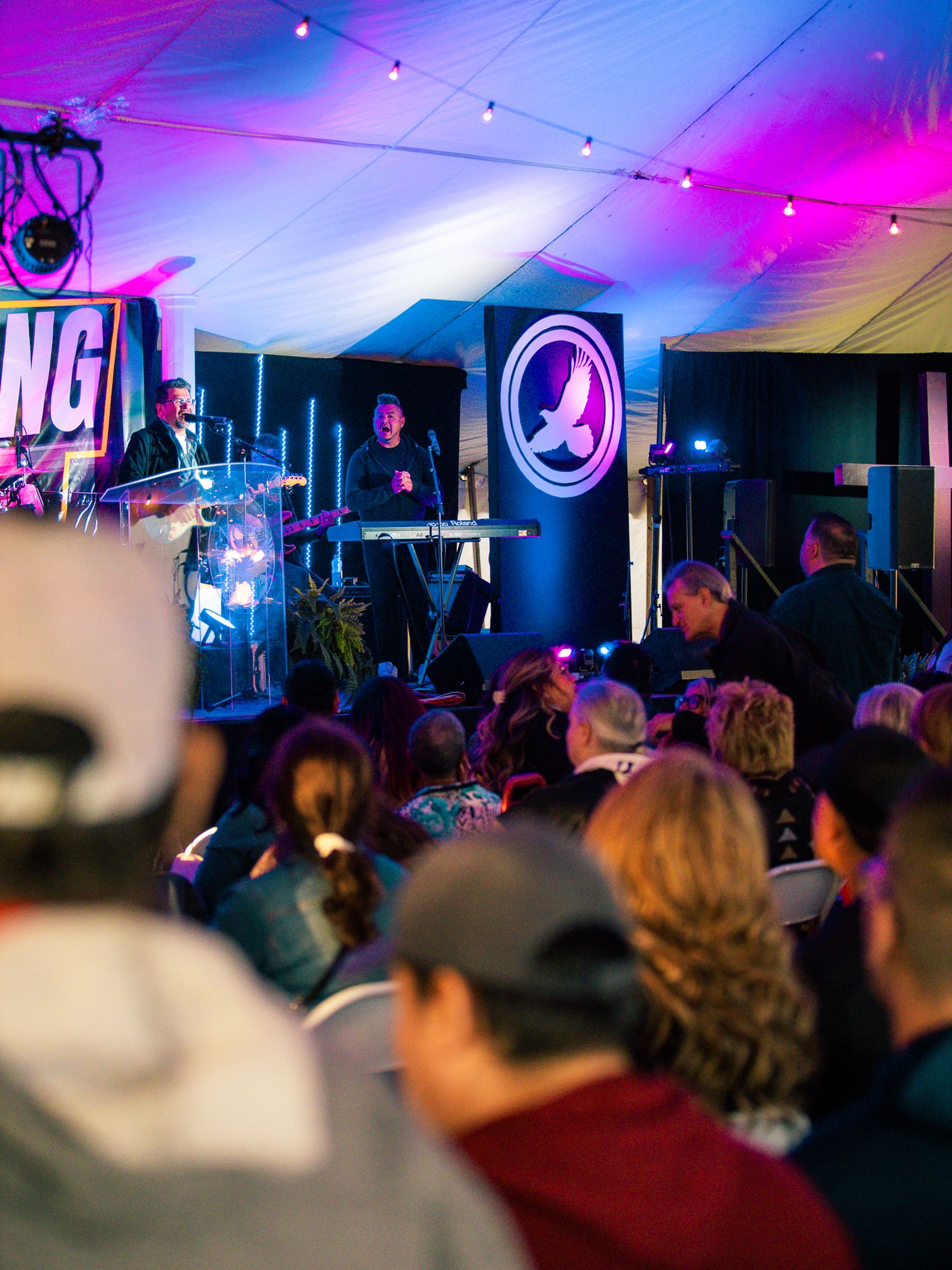 Crowd gathered at a vibrant evening event under a tent with colorful purple lighting and a speaker at the podium.