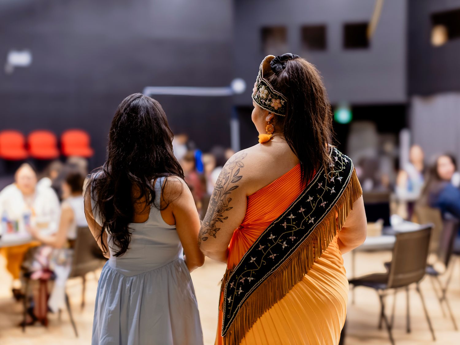 Two women in traditional Indigenous attire stand together in a busy indoor event venue.