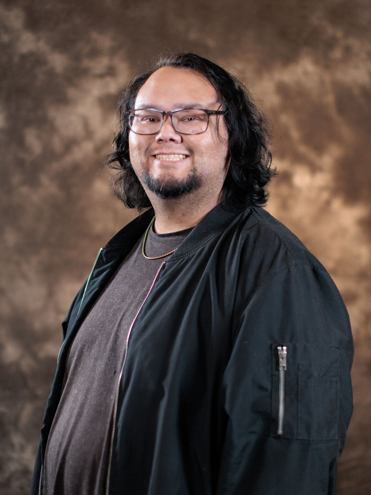 Smiling man with glasses and long hair wearing black bomber jacket in studio portrait.