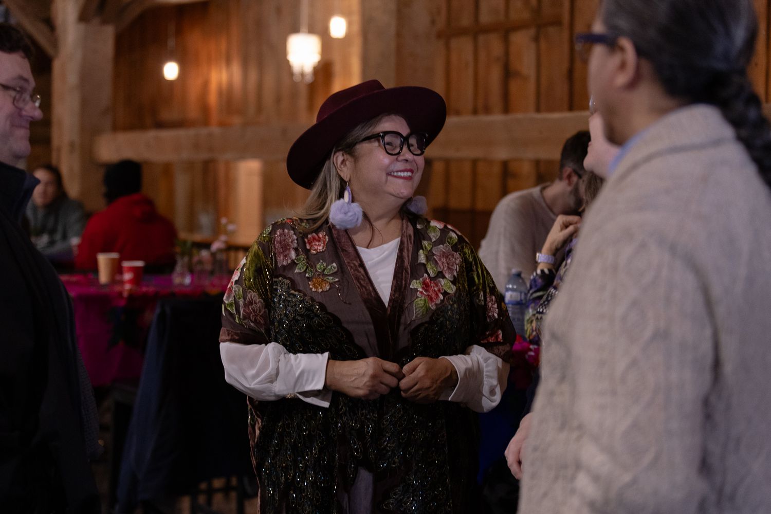 Woman in floral vest and burgundy hat smiling at a social gathering inside a warmly lit barn venue.