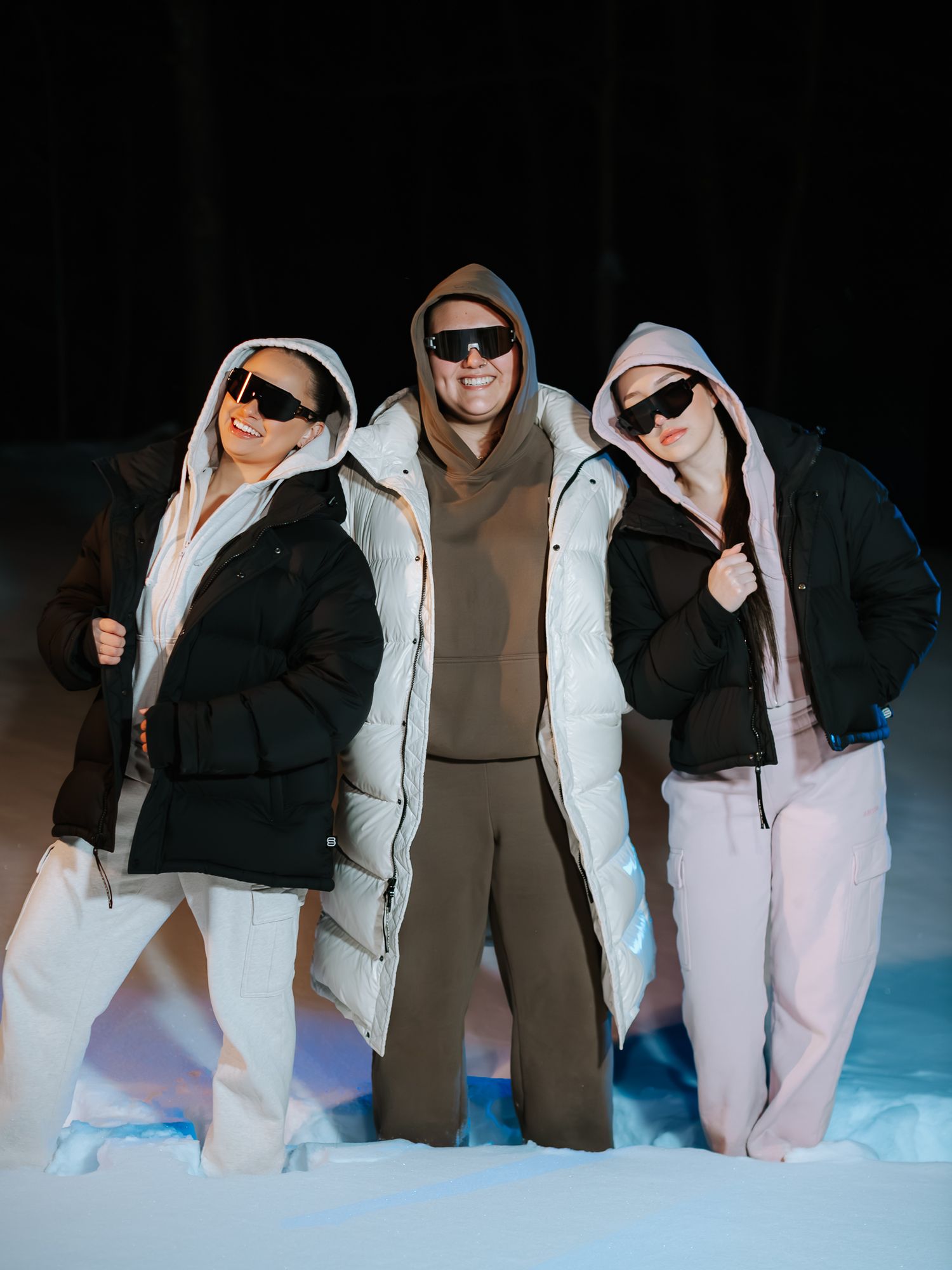 Three models in hooded puffer jackets and sunglasses posing confidently in snow during a winter night shoot.