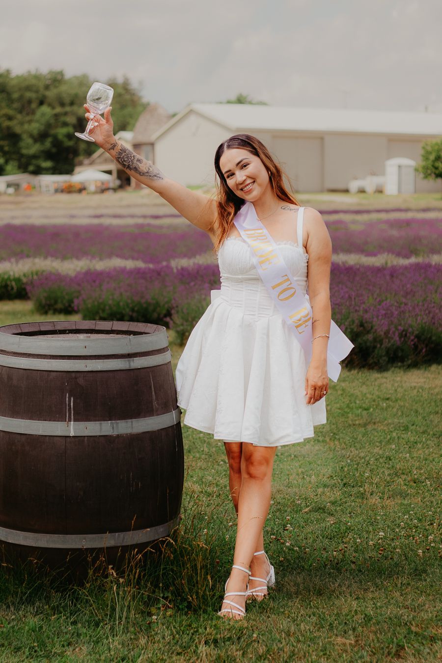 Woman in white dress posing with wine glass near wooden barrel in lavender field.