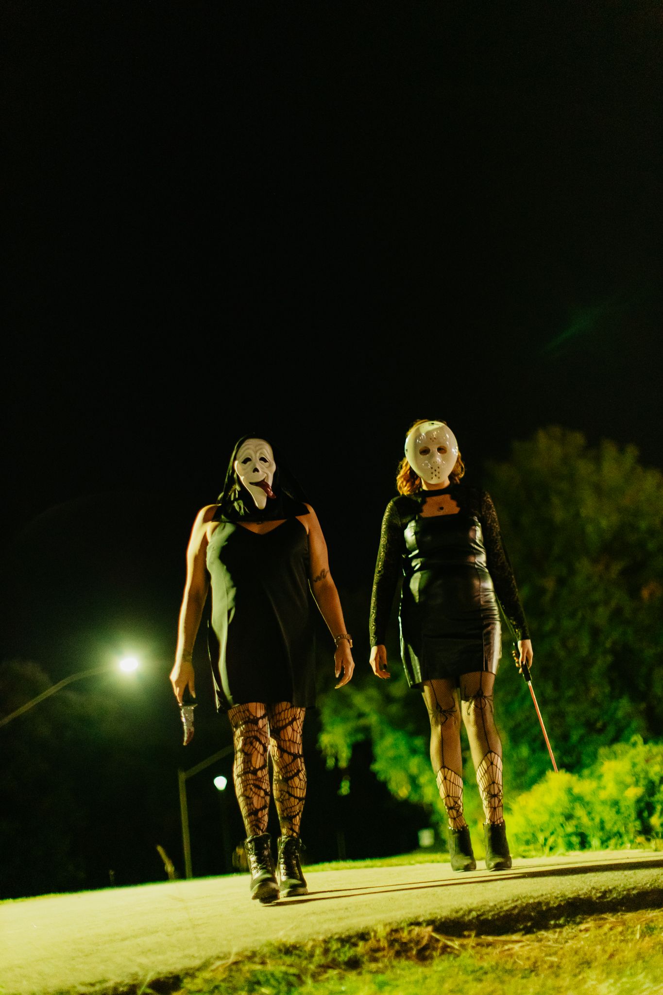 Two figures in white masks and dark clothing walking on dimly lit path at night.