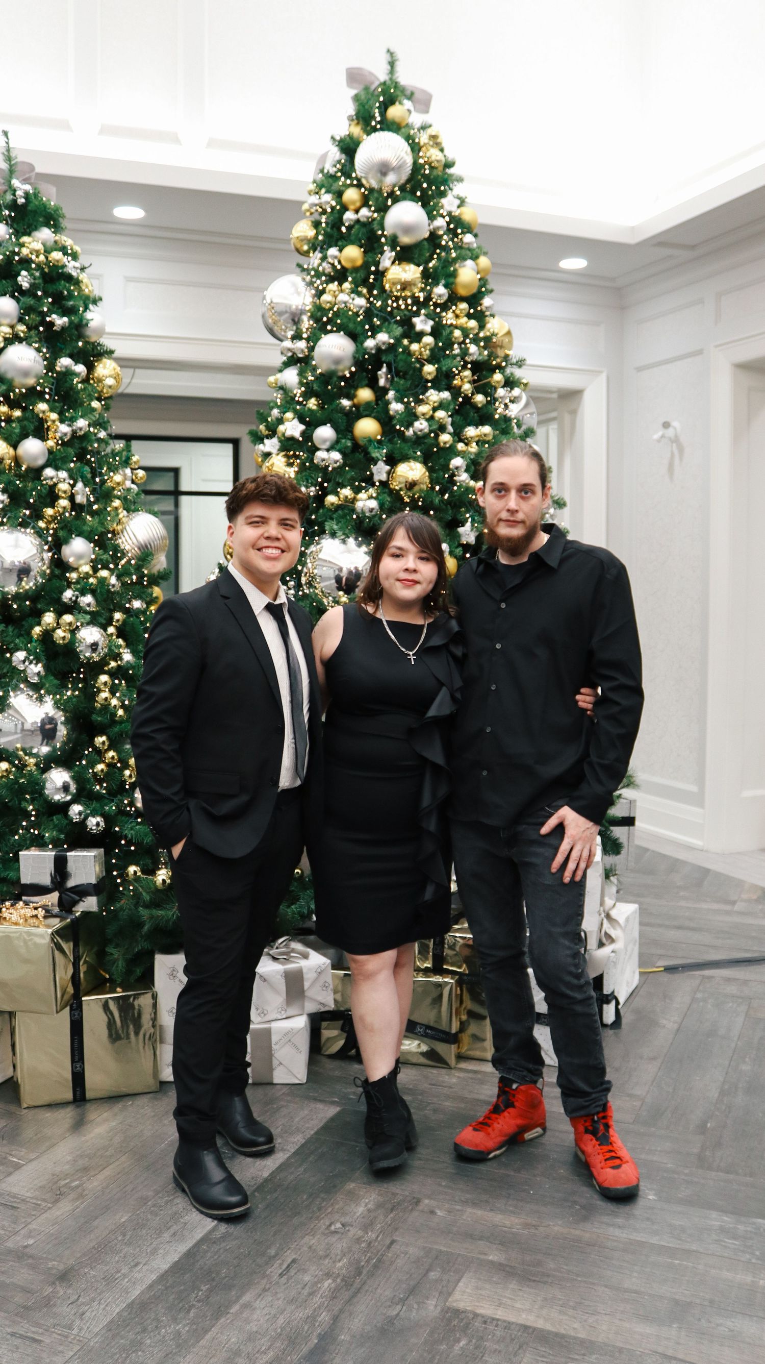 Three people in formal black attire pose in front of a decorated Christmas tree in an elegant white room.