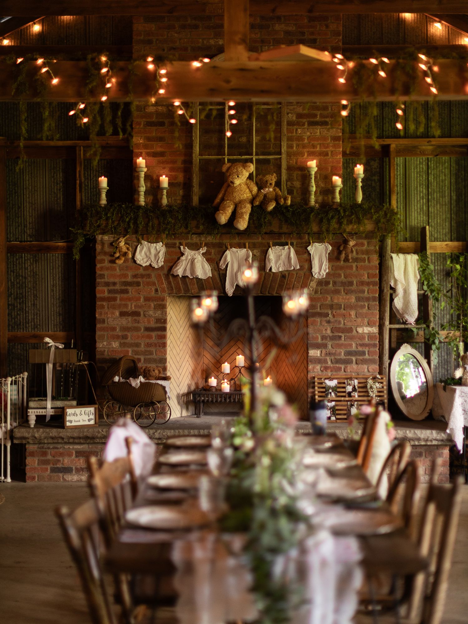 A rustic barn reception setup with string lights, baby clothes on display, and teddy bears decorating the exposed brick walls.