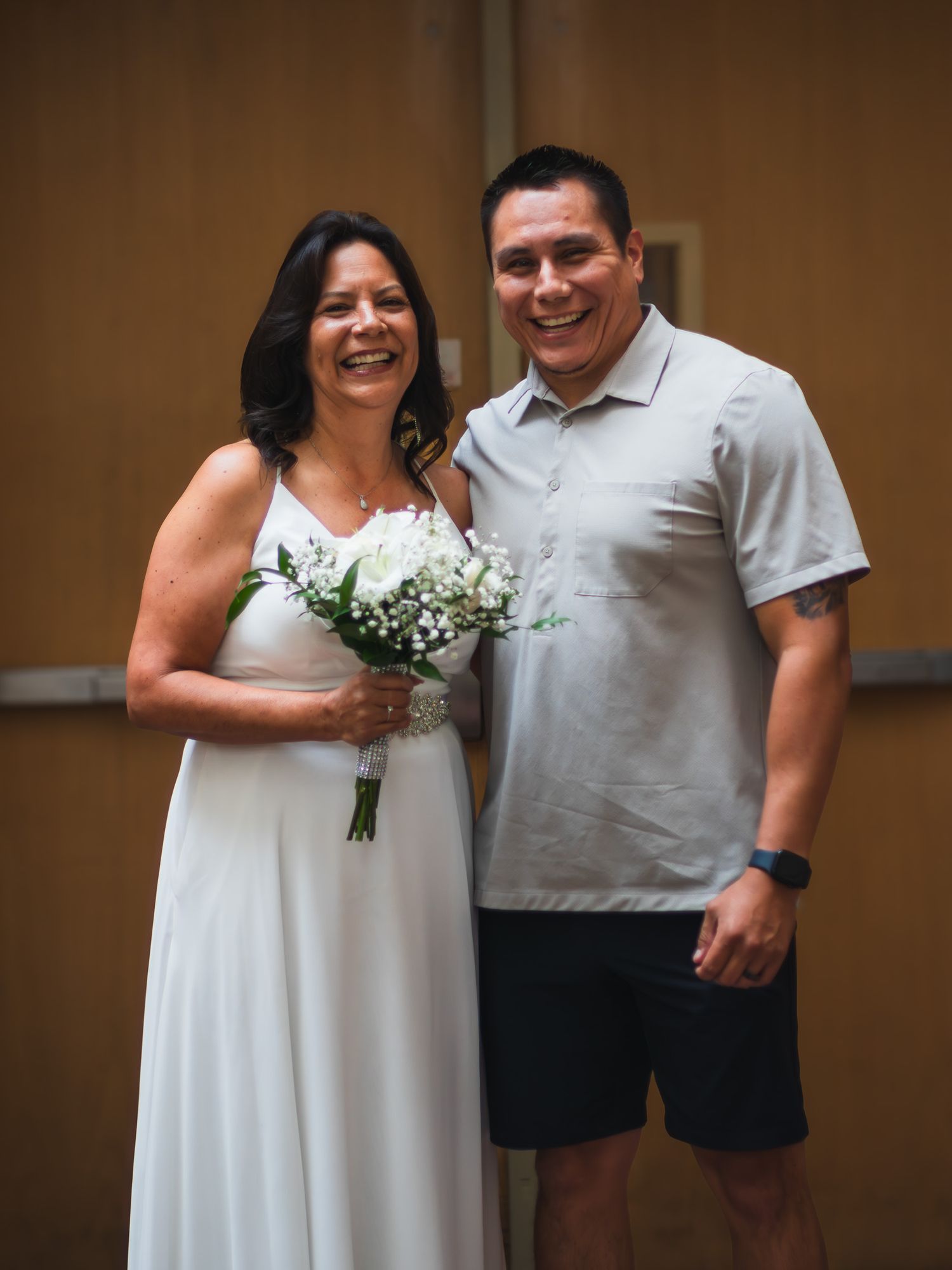 Smiling bride in white halter gown holds white bouquet beside a man in a gray polo shirt at a wedding celebration.
