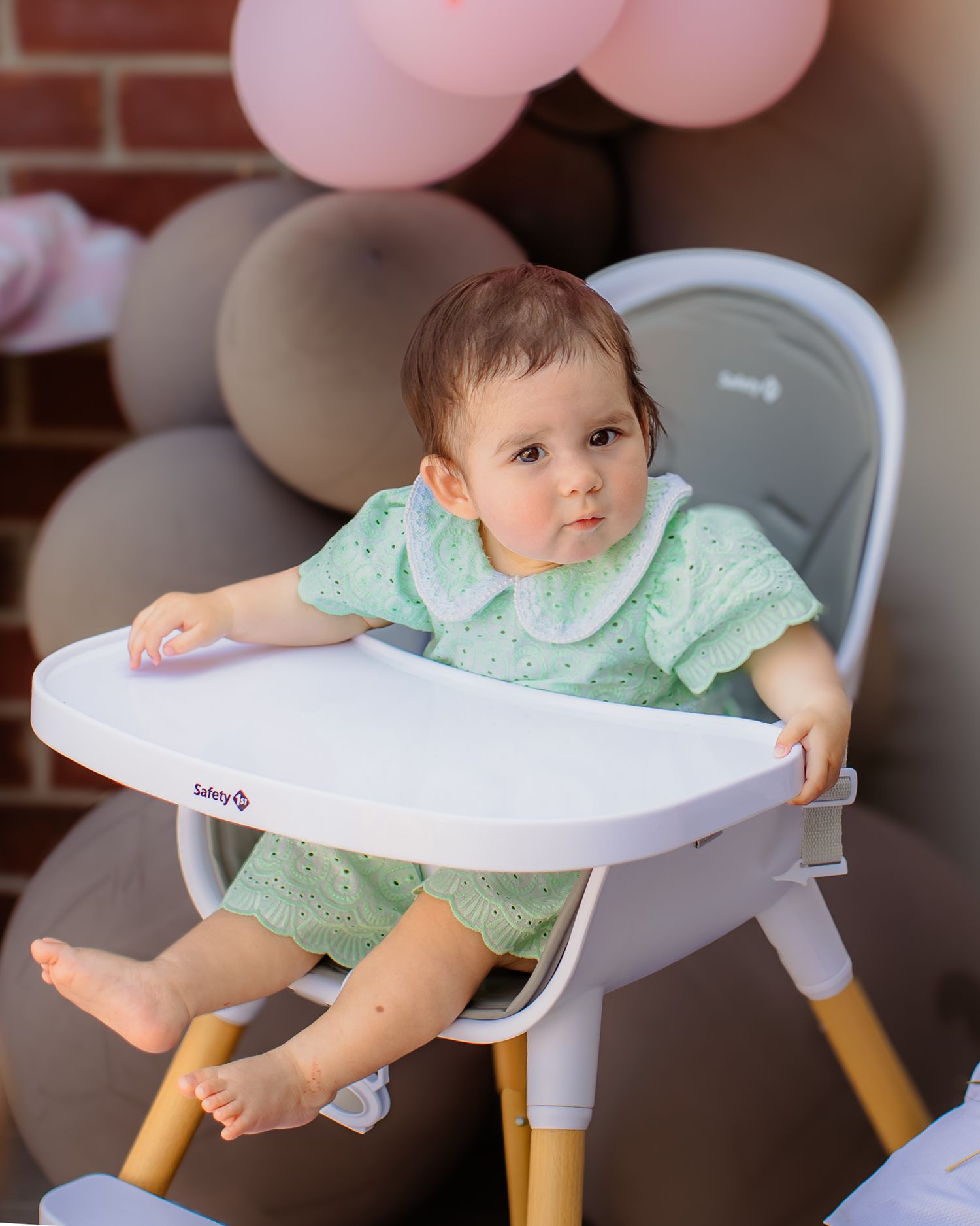 Baby girl in mint green outfit sits in a Safety 1st high chair surrounded by pink balloons at a first birthday party.