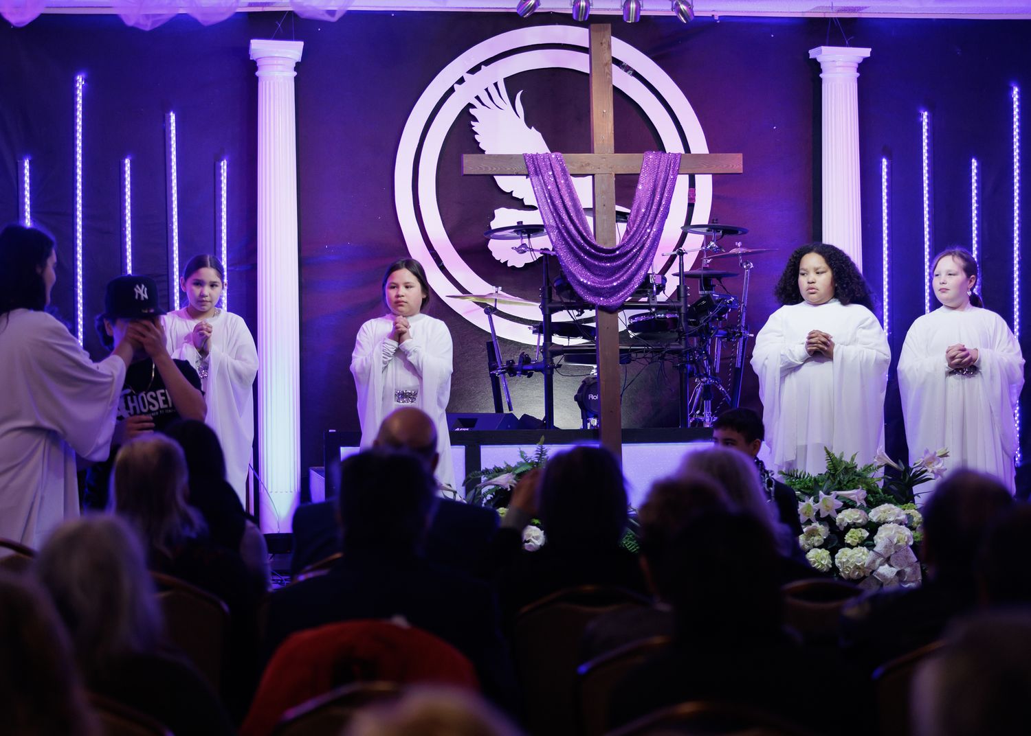 Young women in white robes perform on a church stage with a purple-lit cross and dove symbol during a religious service.