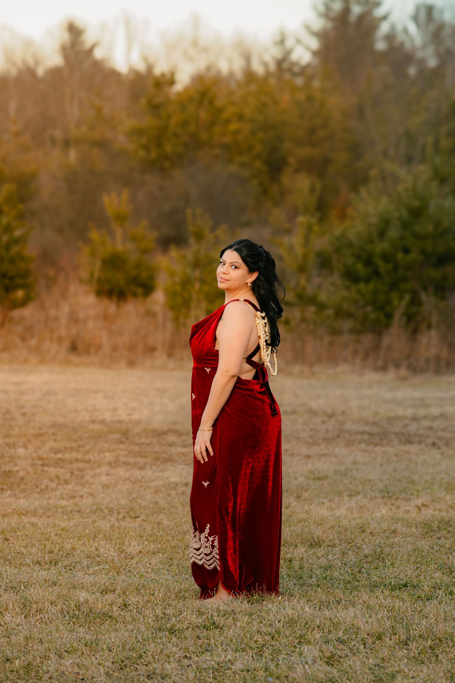 Woman in elegant red velvet saree posing in golden hour light in open field.