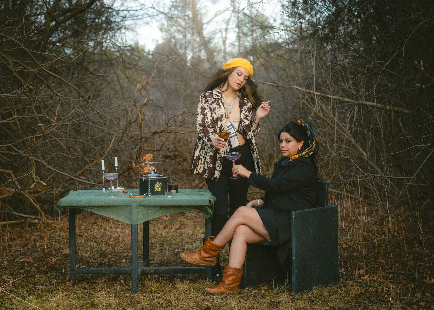 Two women in bohemian outfits sit and stand at a vintage green table outdoors in a wooded setting.