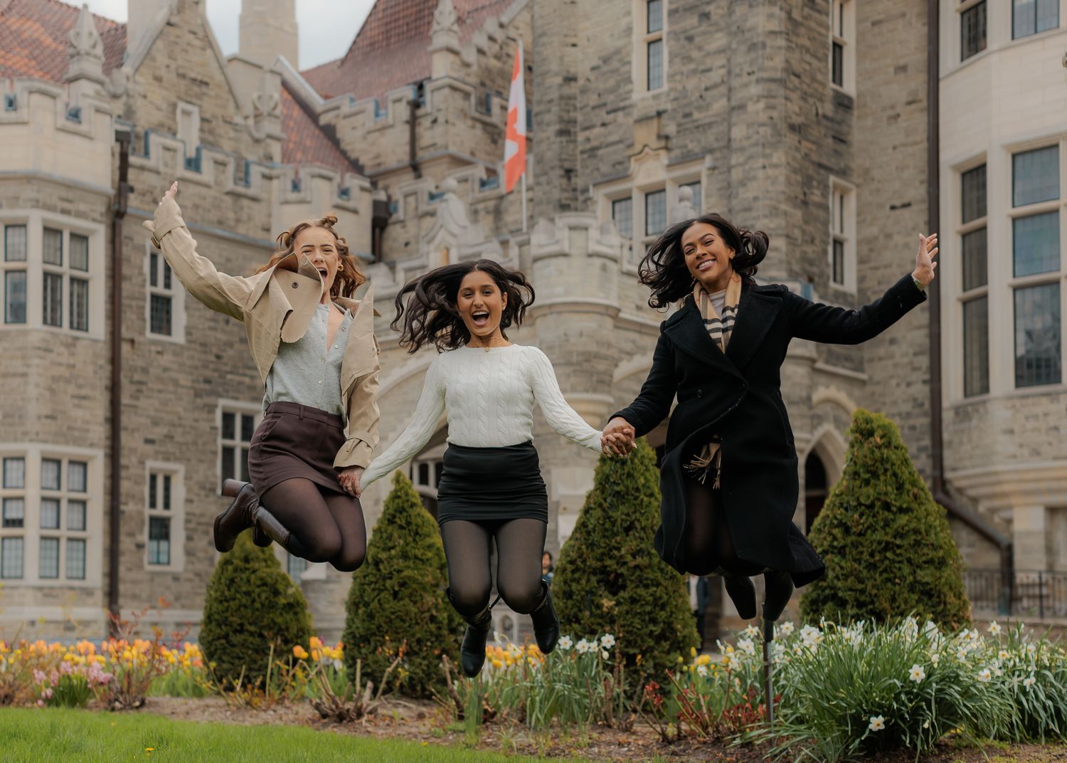 Three joyful women jumping in front of a historic castle building surrounded by spring flowers and greenery.