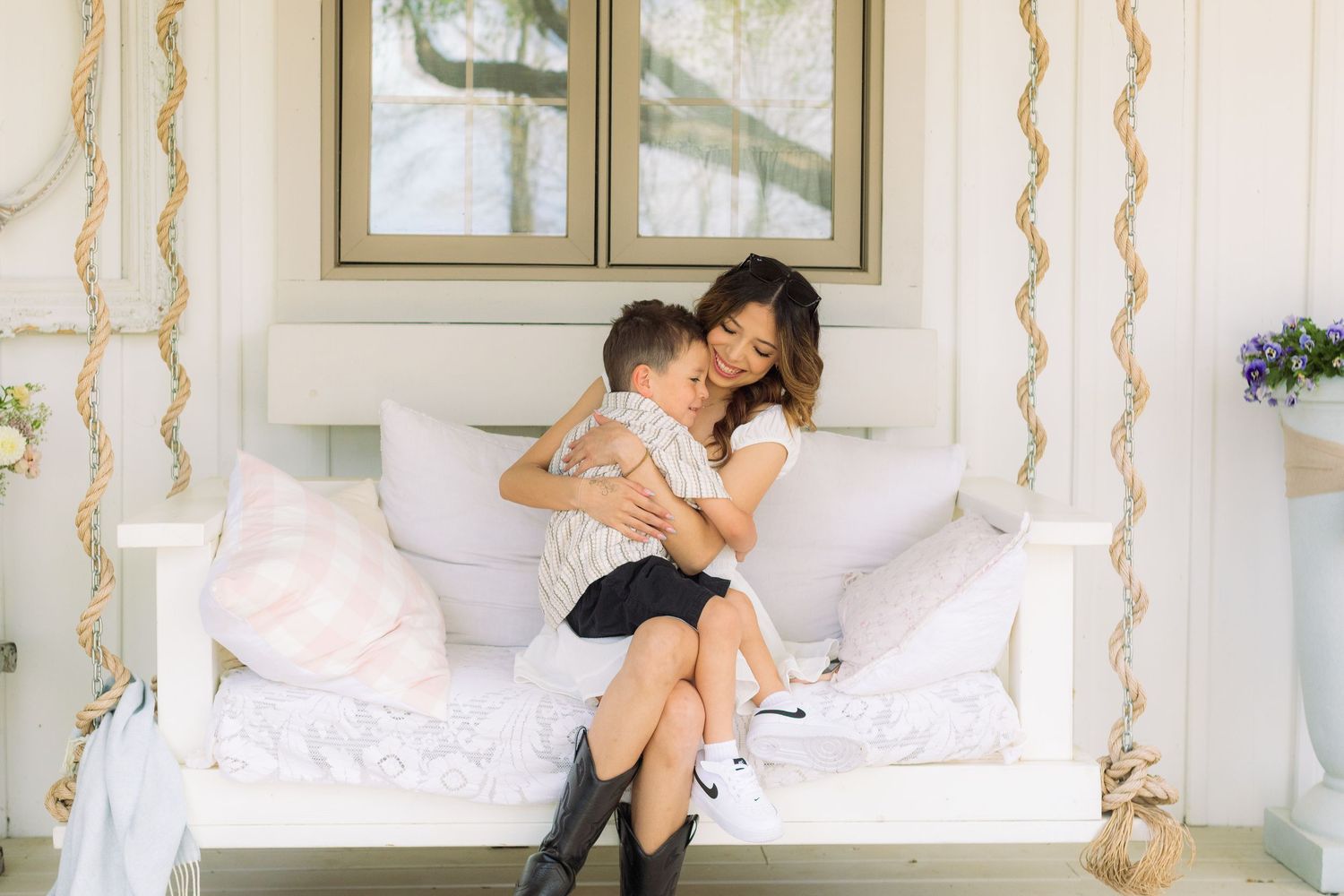 A mother and young son share a tender embrace on a white porch swing adorned with rope accents and pink pillows.