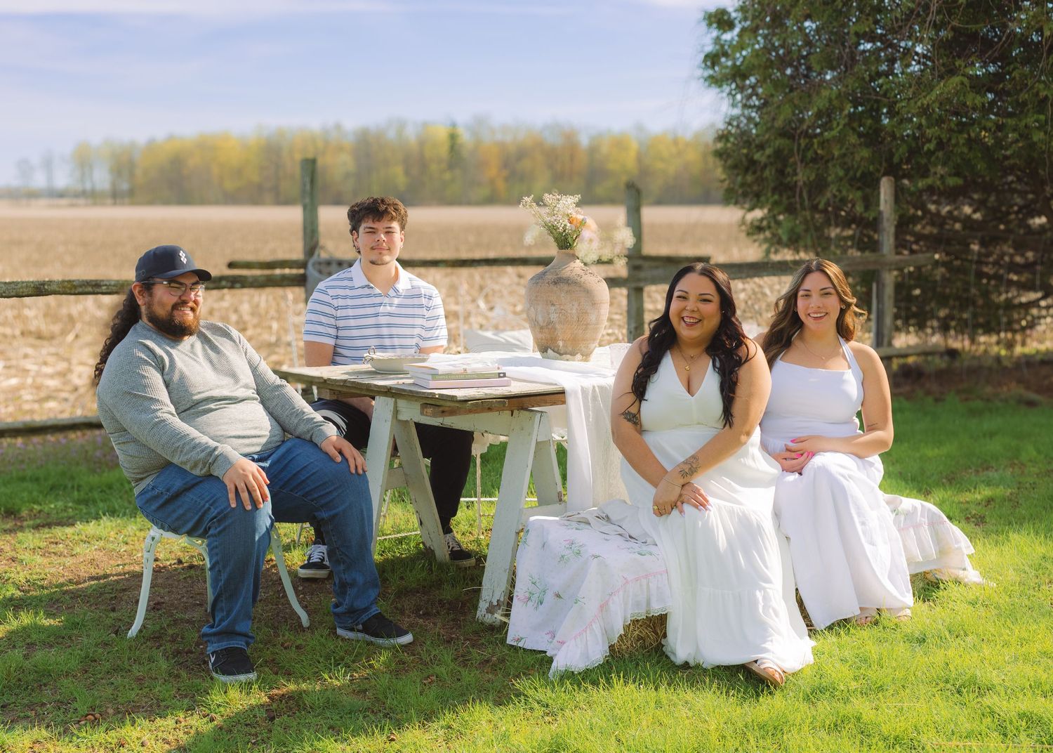 Four people sitting outdoors at a white table on a green lawn with a wooden fence and trees in the background.