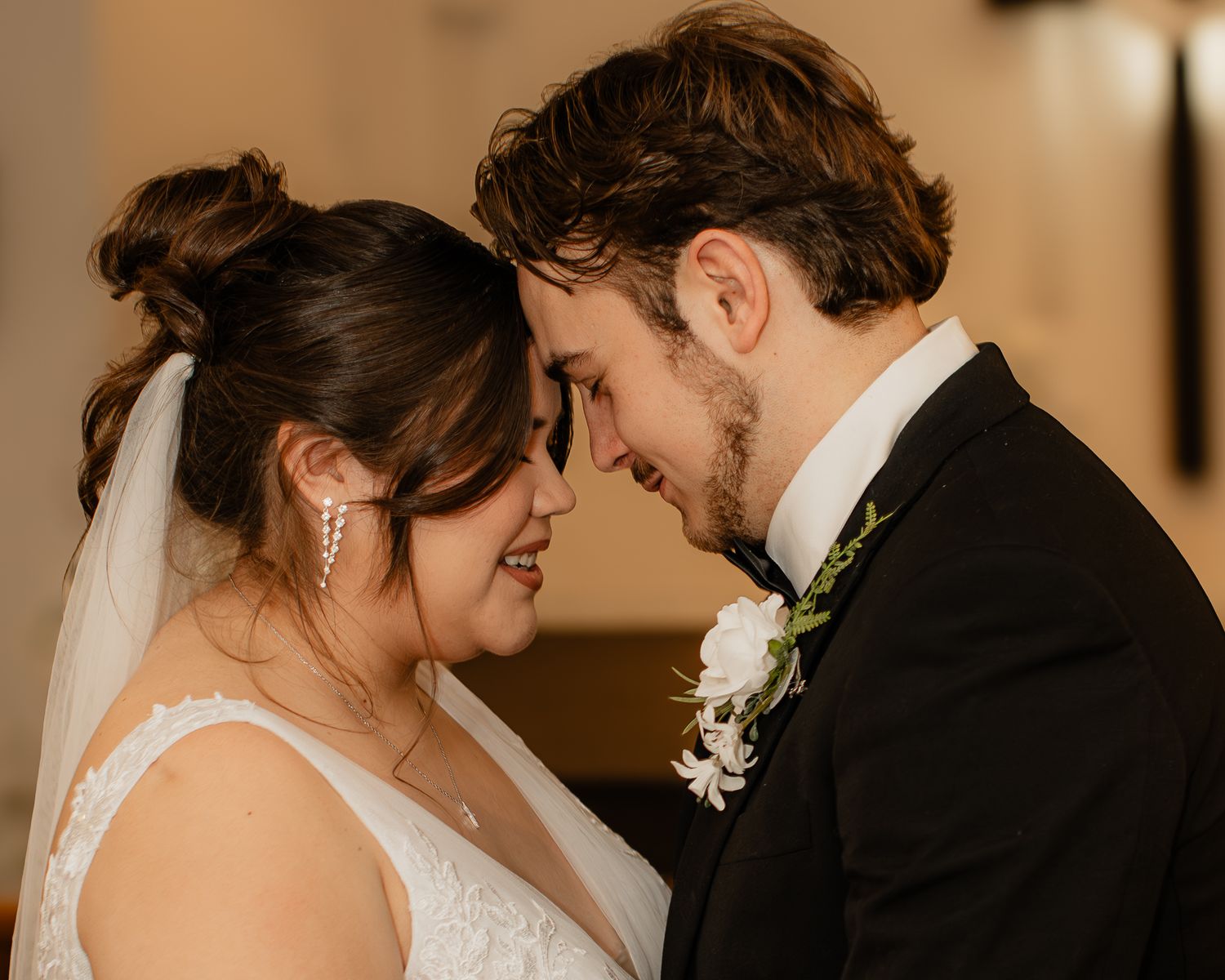 A bride and groom share an intimate forehead touch moment, smiling softly in wedding attire with veil and boutonniere.