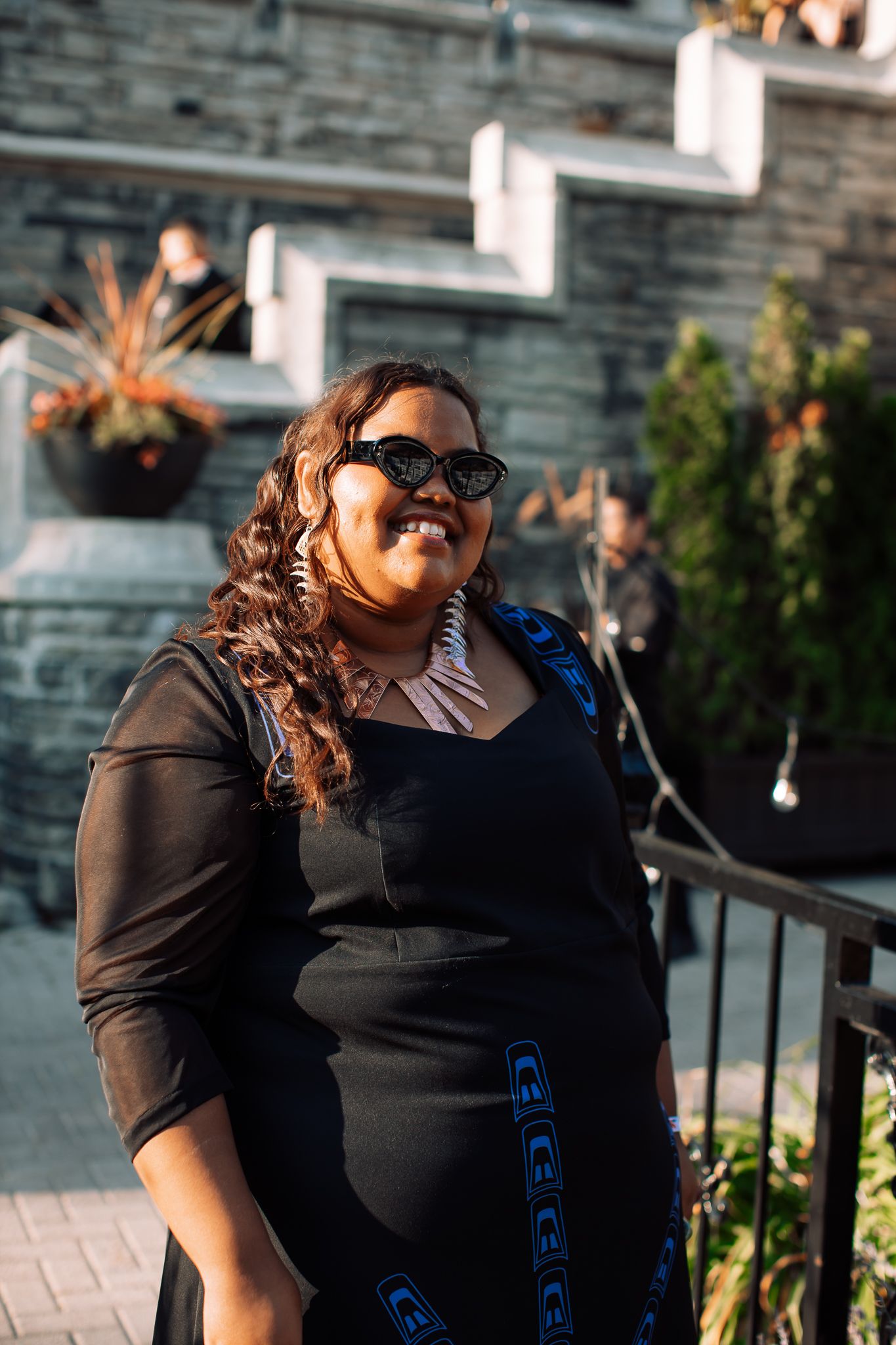 Smiling plus-size woman wearing sunglasses and black dress with blue embroidery outdoors.
