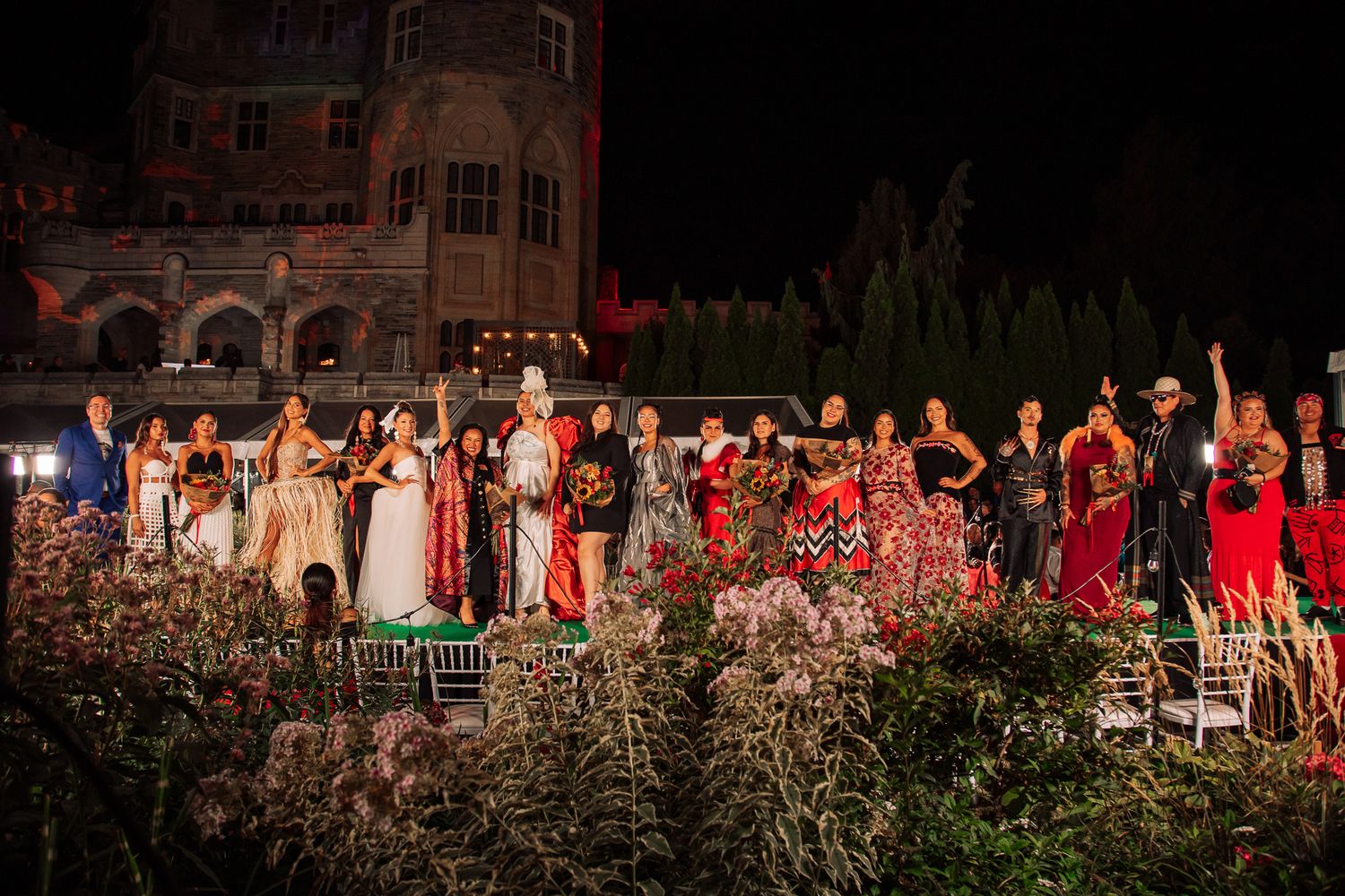 Large group of diverse models in colorful traditional attire pose on an outdoor stage at night before a historic building.