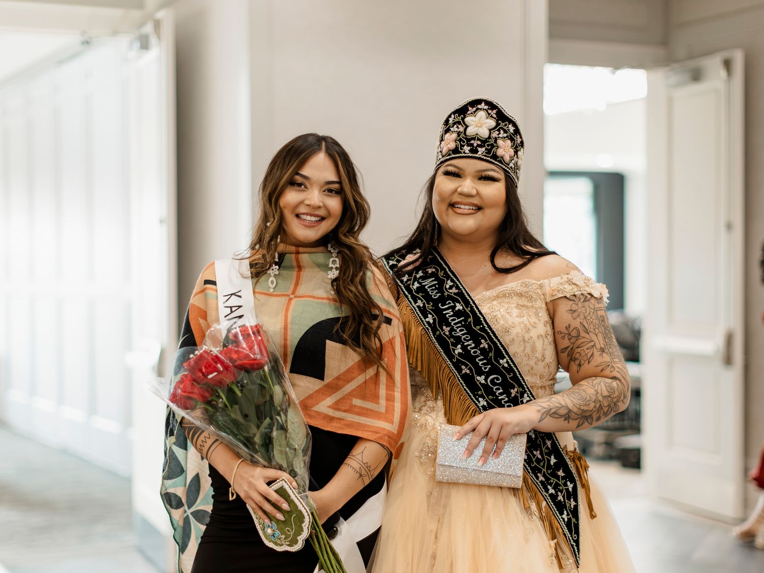 Two smiling women pose together at a pageant, one wearing a sash and holding red roses, one in a crown.
