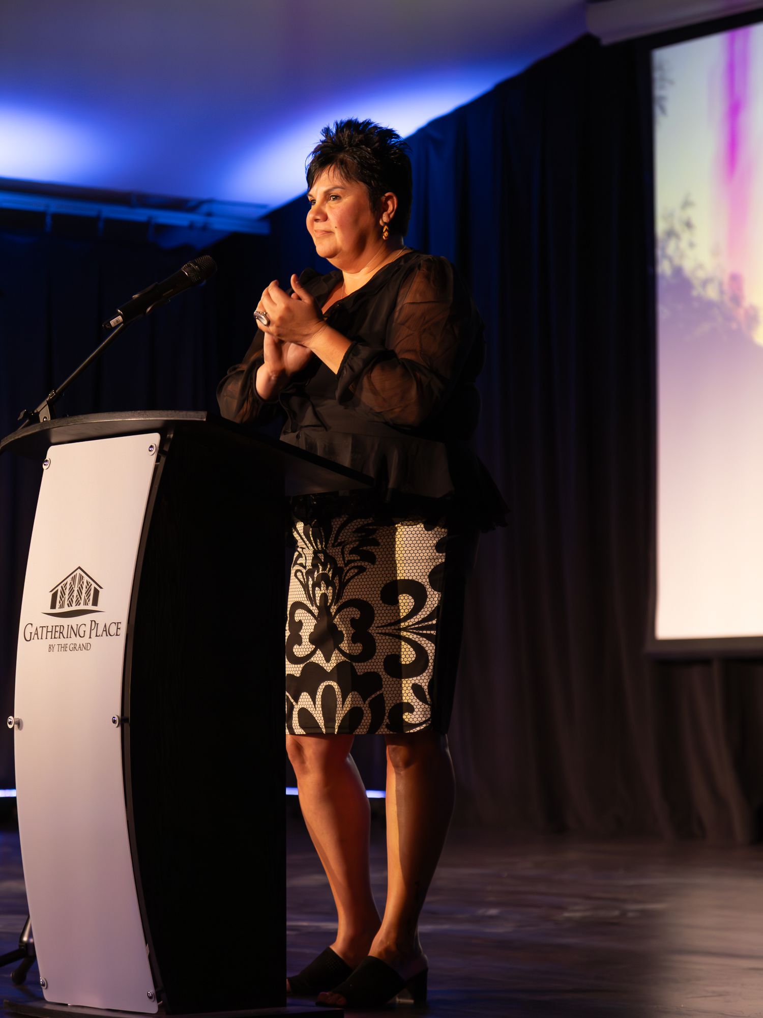 Woman in black outfit speaks at podium at Gathering Place at The Sand event venue with blue stage lighting.
