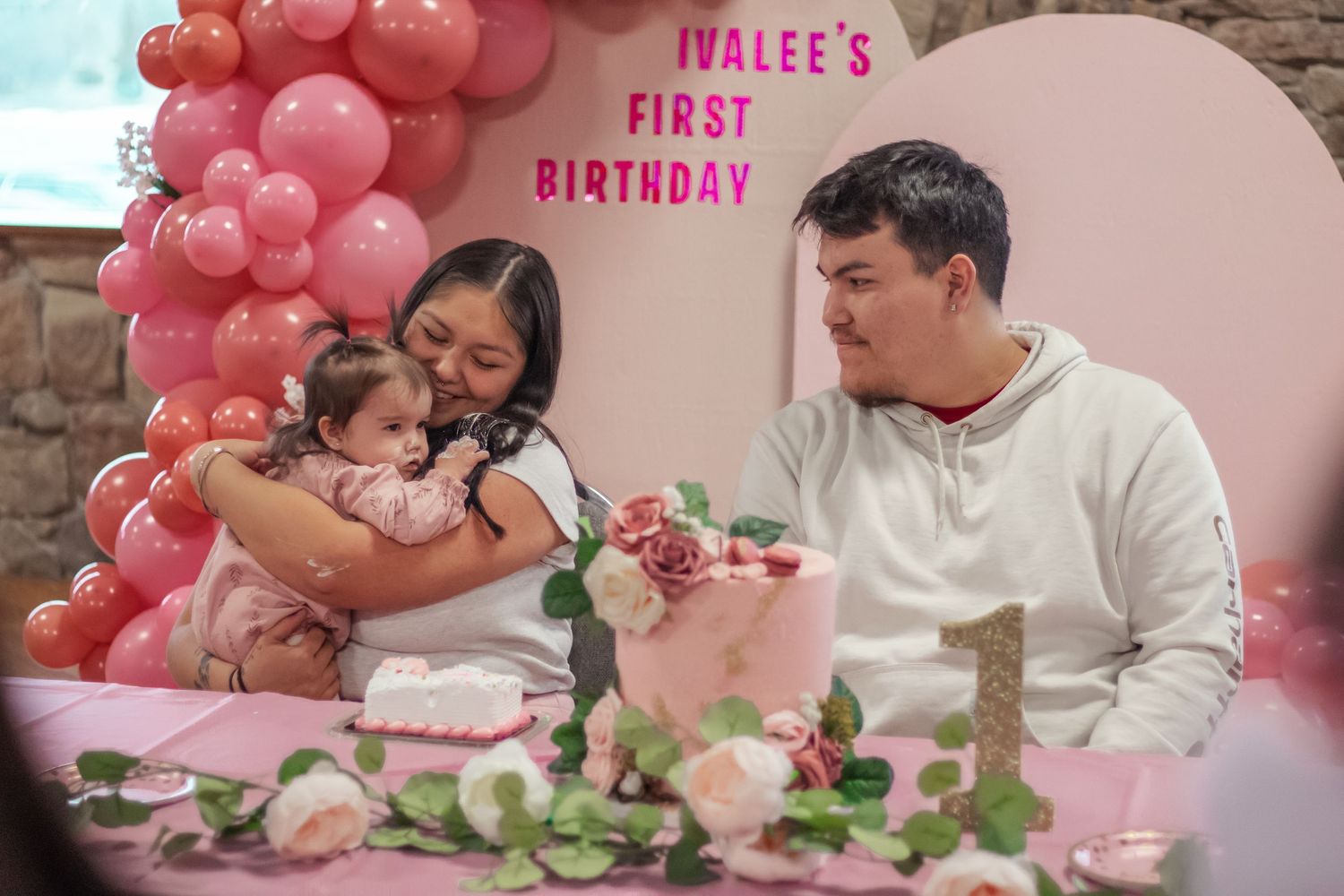 Family celebrating baby Ivalee's first birthday with pink balloons, floral cake, and decorations.