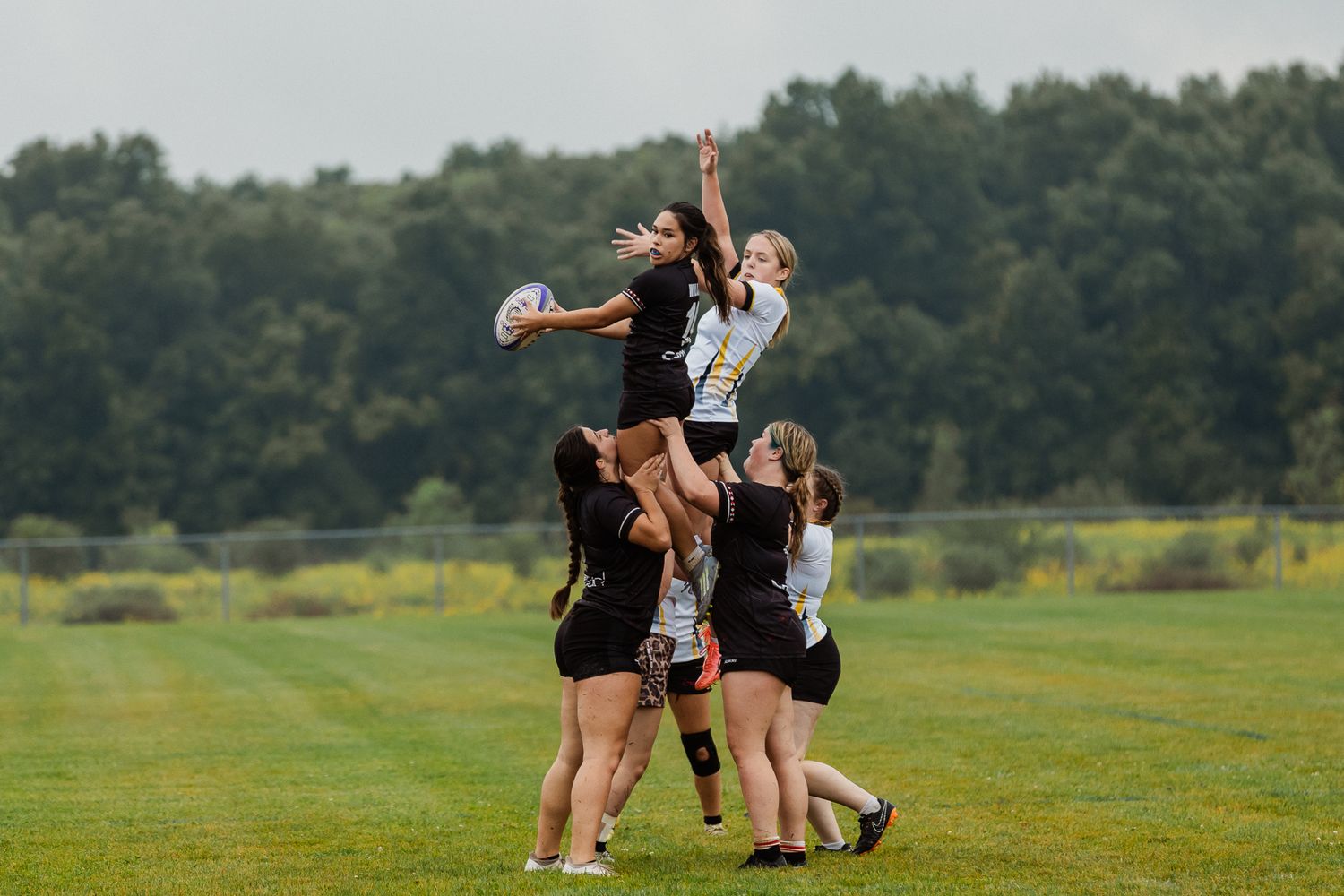 Women's rugby lineout with players lifting teammate to catch ball during outdoor match on green field.