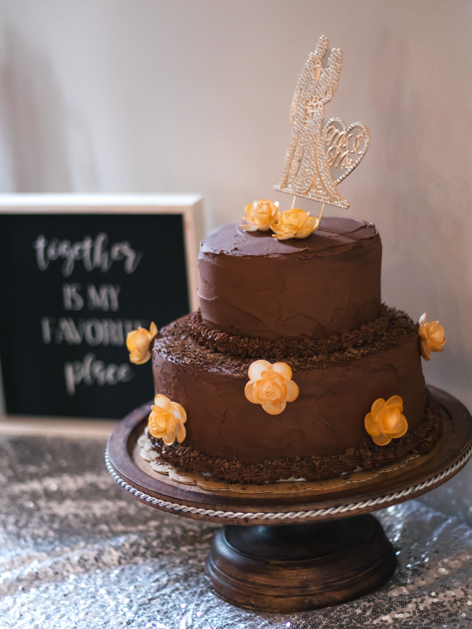 Two-tier chocolate wedding cake with gold floral topper and yellow roses next to together is my favorite place sign.