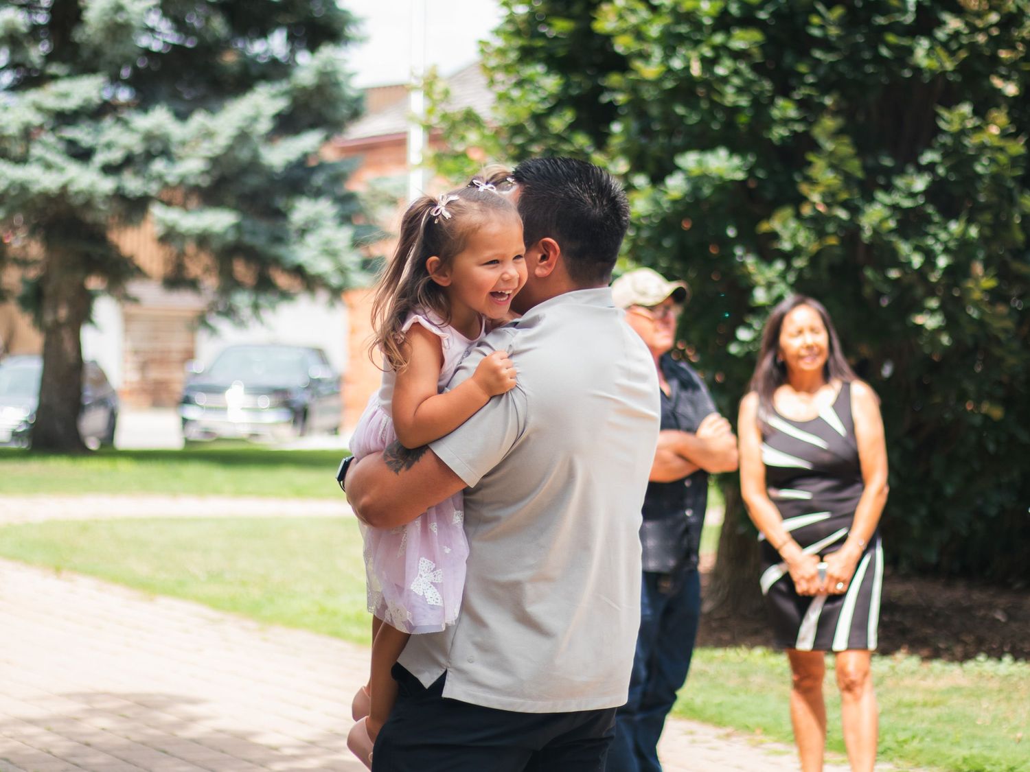 A man joyfully hugs a laughing little girl outdoors, with a woman smiling in the background on a sunny day.