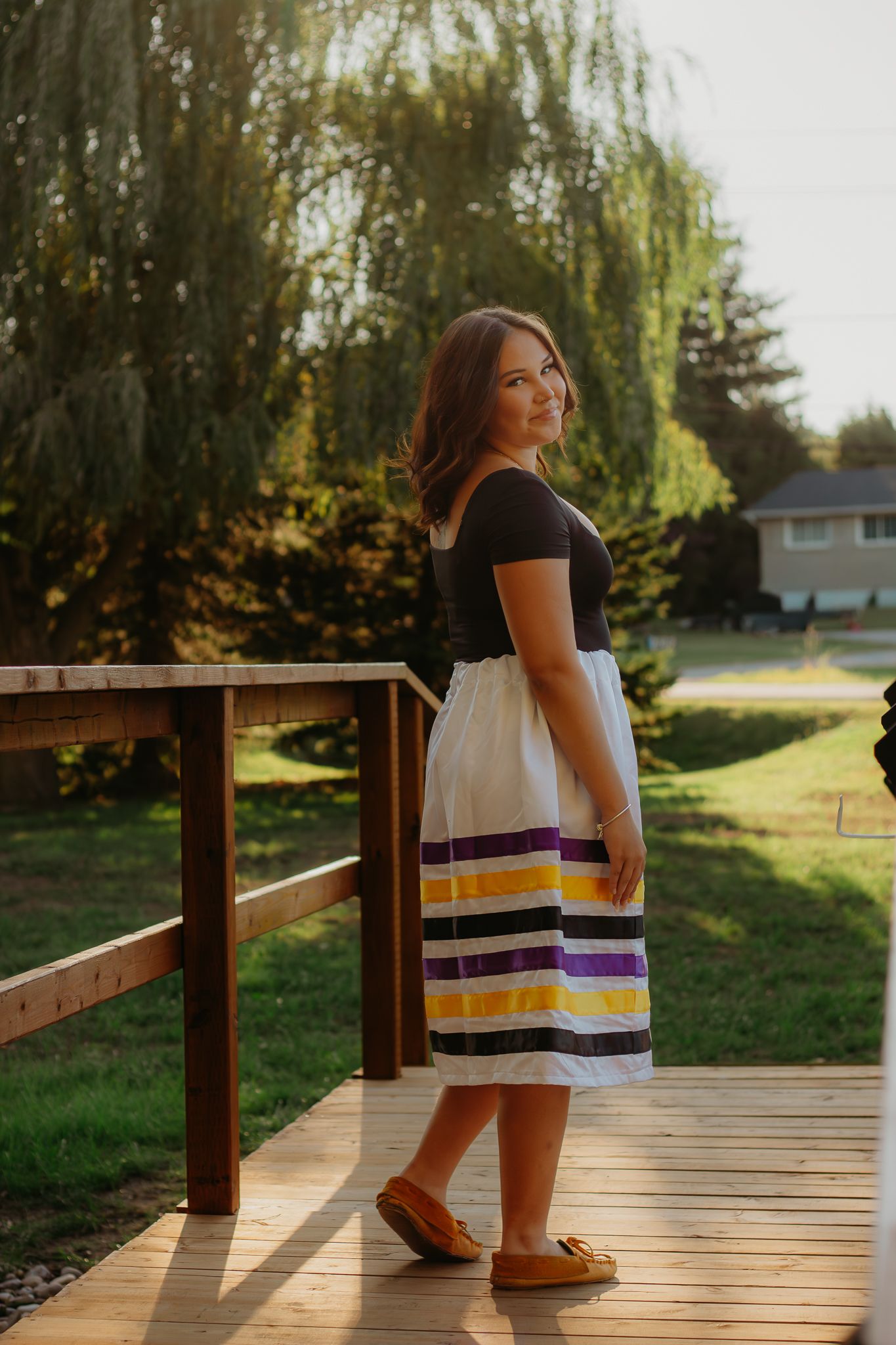Graduate in striped dress poses on wooden boardwalk during sunset.