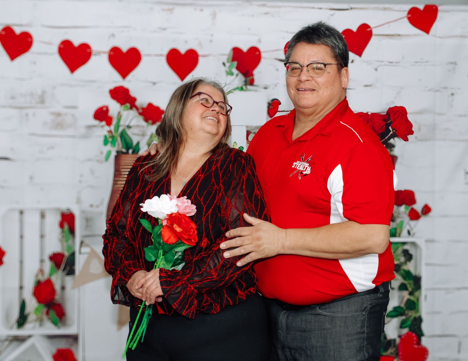 Couple poses against white brick wall decorated with red hearts and roses for Valentine's Day.