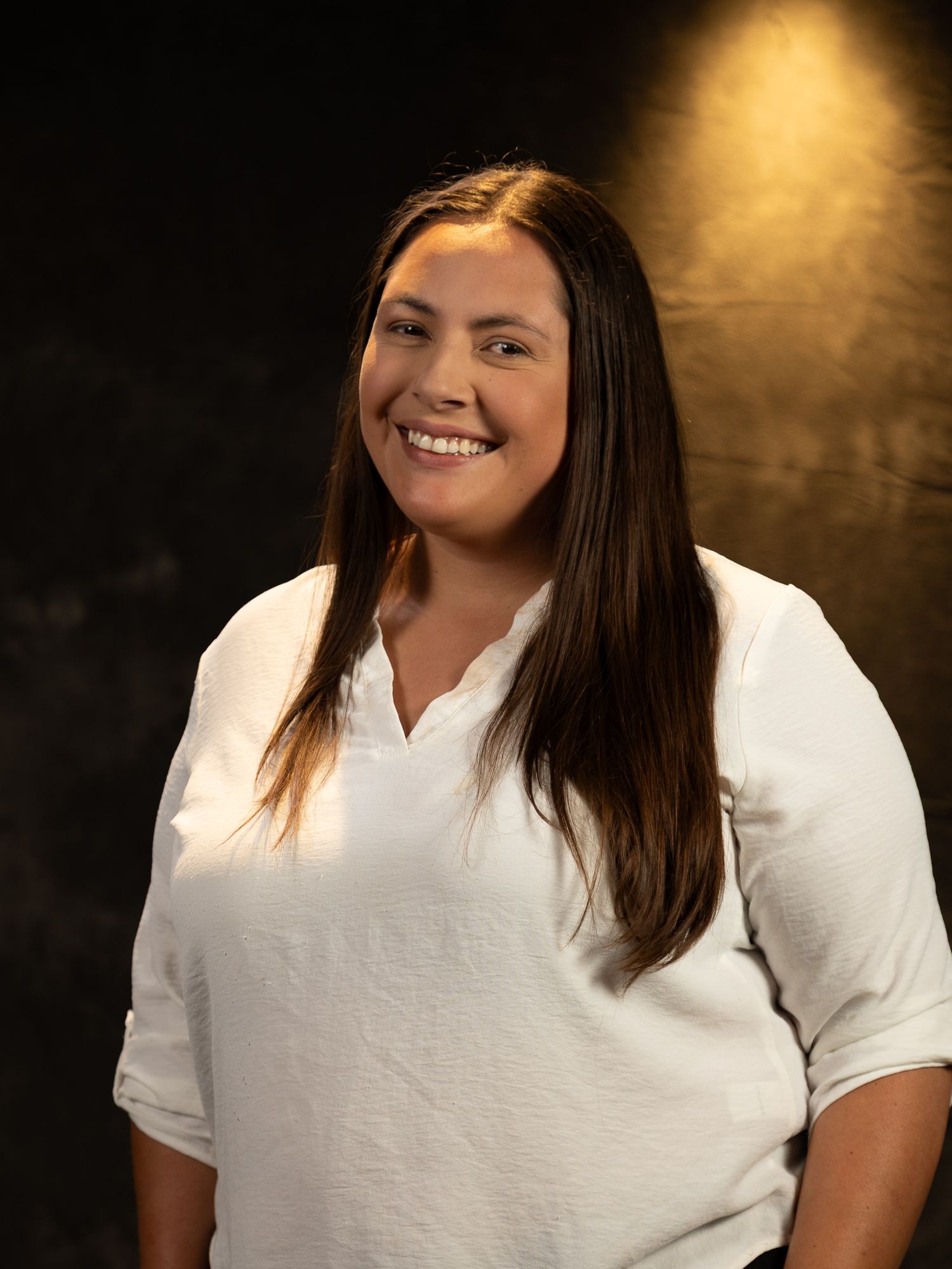 Professional headshot of person in white top with warm lighting against dark background.