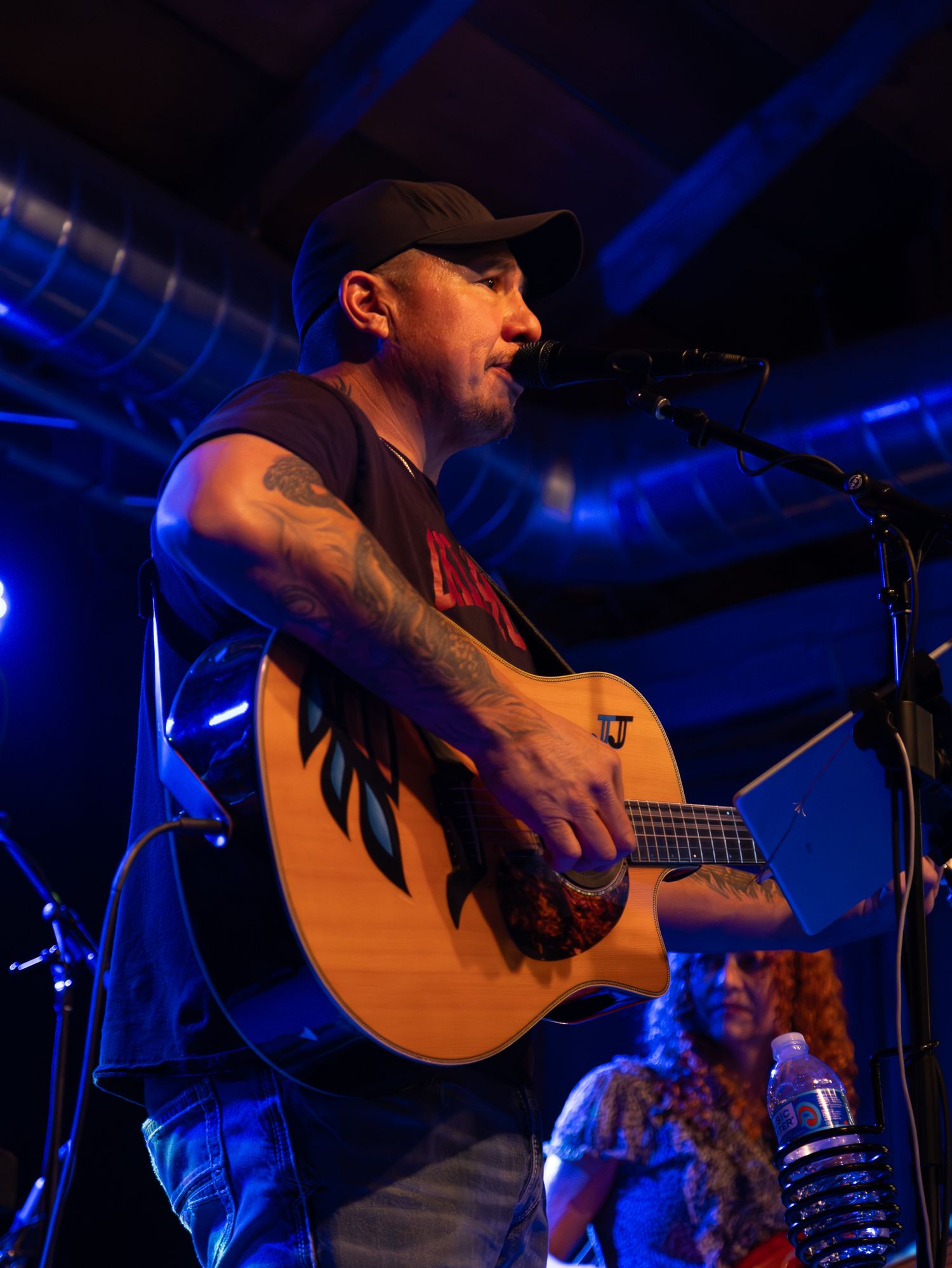 A musician in a baseball cap plays acoustic guitar under blue stage lighting.