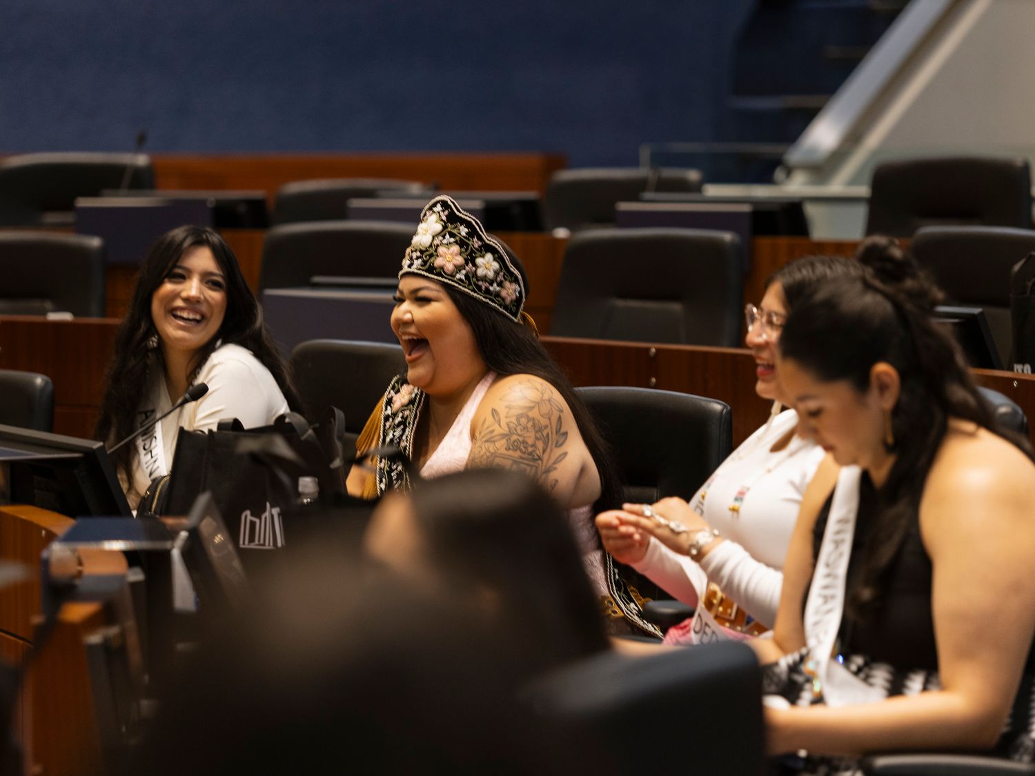 Indigenous women in traditional regalia laugh together in an auditorium setting.