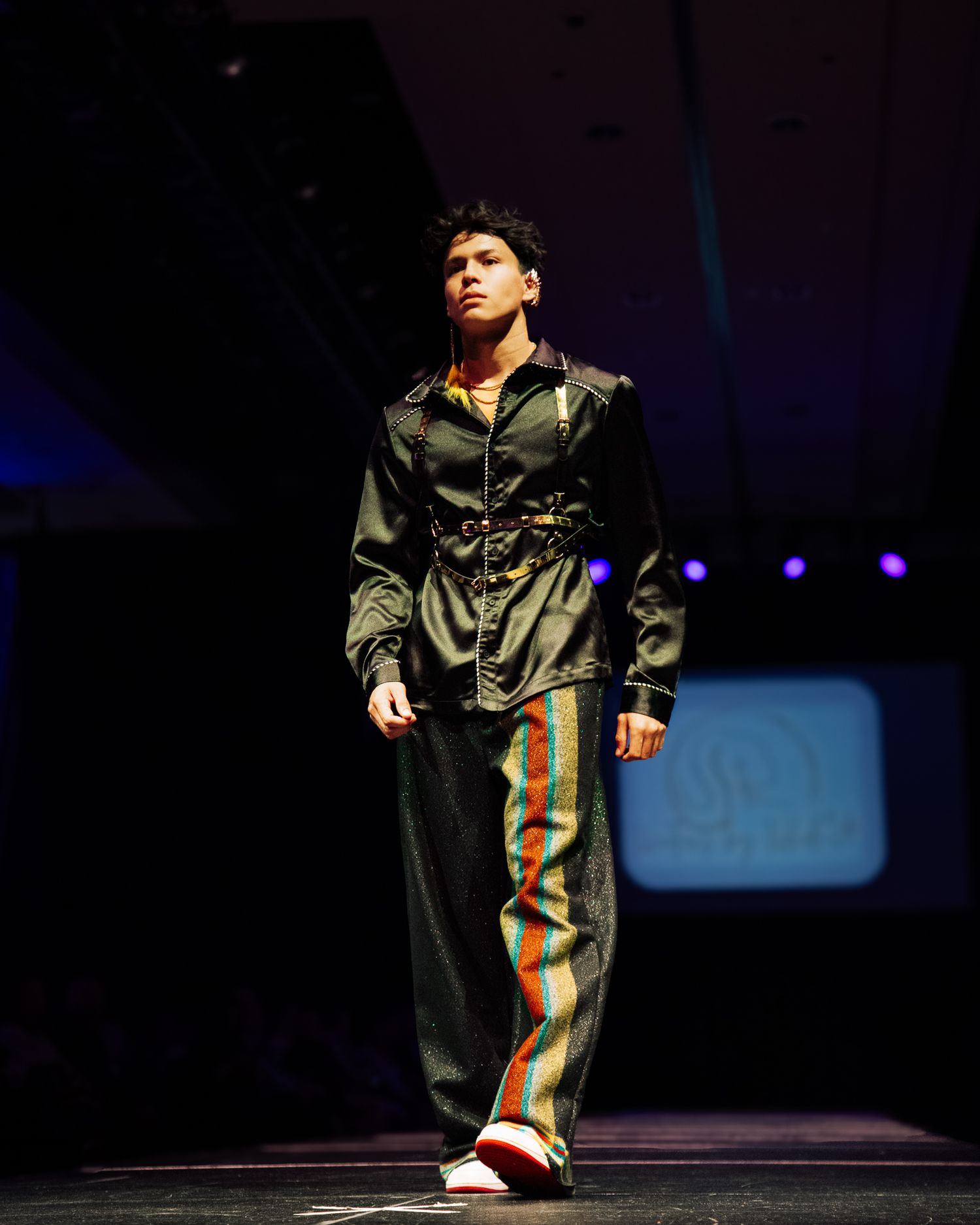 Model walks runway in dark satin jacket, striped pants, and colorful sneakers at a fashion show.
