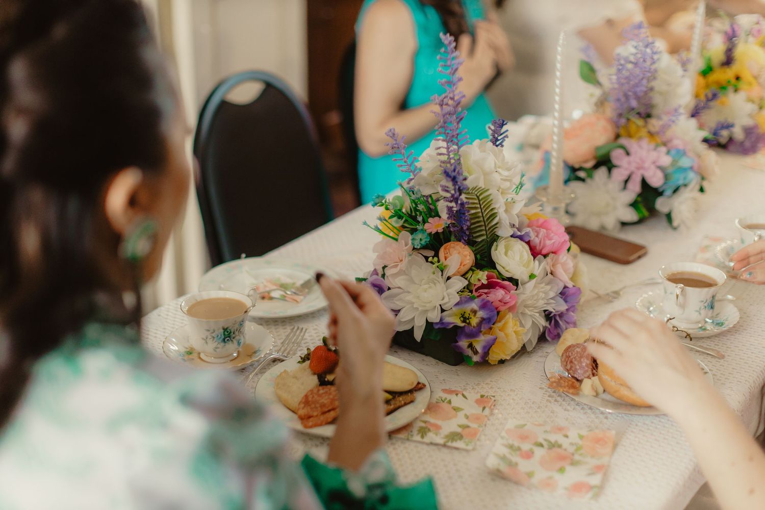 Women in teal dresses enjoy a floral-decorated tea party table with colorful bouquets and fine china cups.