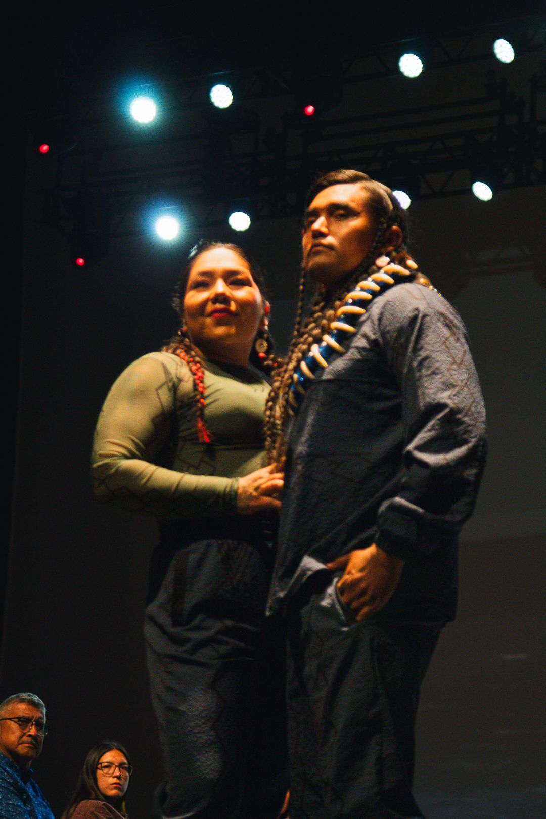 Two models pose together on dark stage wearing traditional Native American braids and contemporary clothing at fashion show.